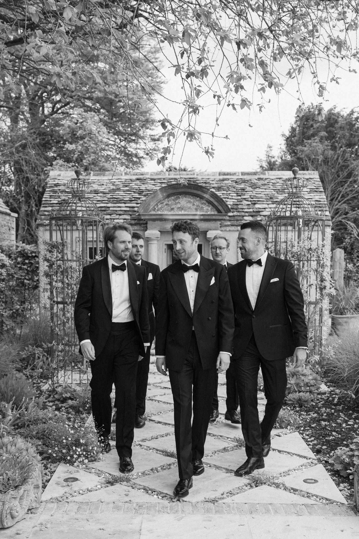 Groomsmen in black tuxedos walking through garden pathway at elegant wedding venue.