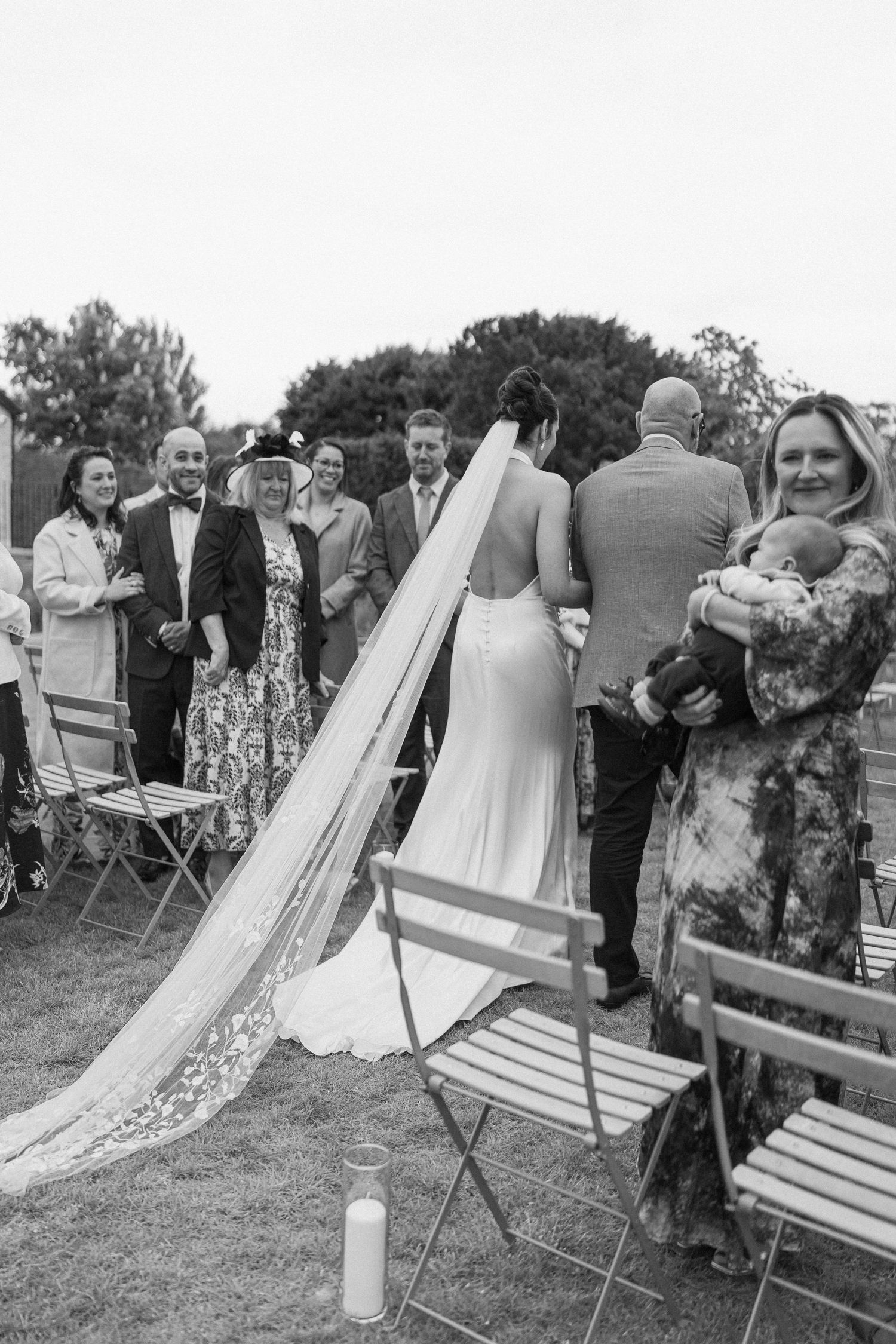 Bride in backless gown with long lace veil walks toward groom at outdoor wedding ceremony with seated guests.