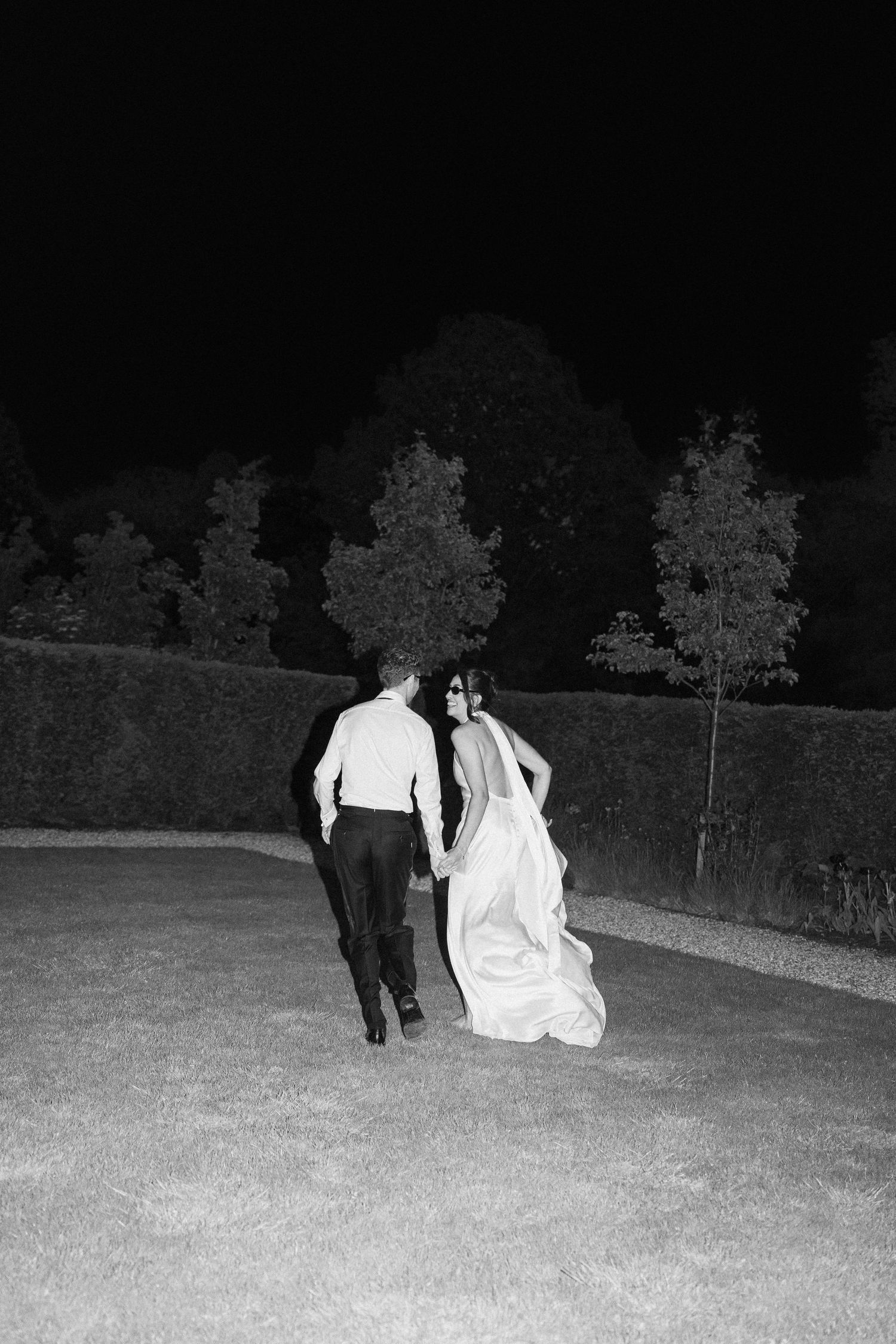 Black and white photo of bride and groom walking hand in hand through dark gardens late at night.