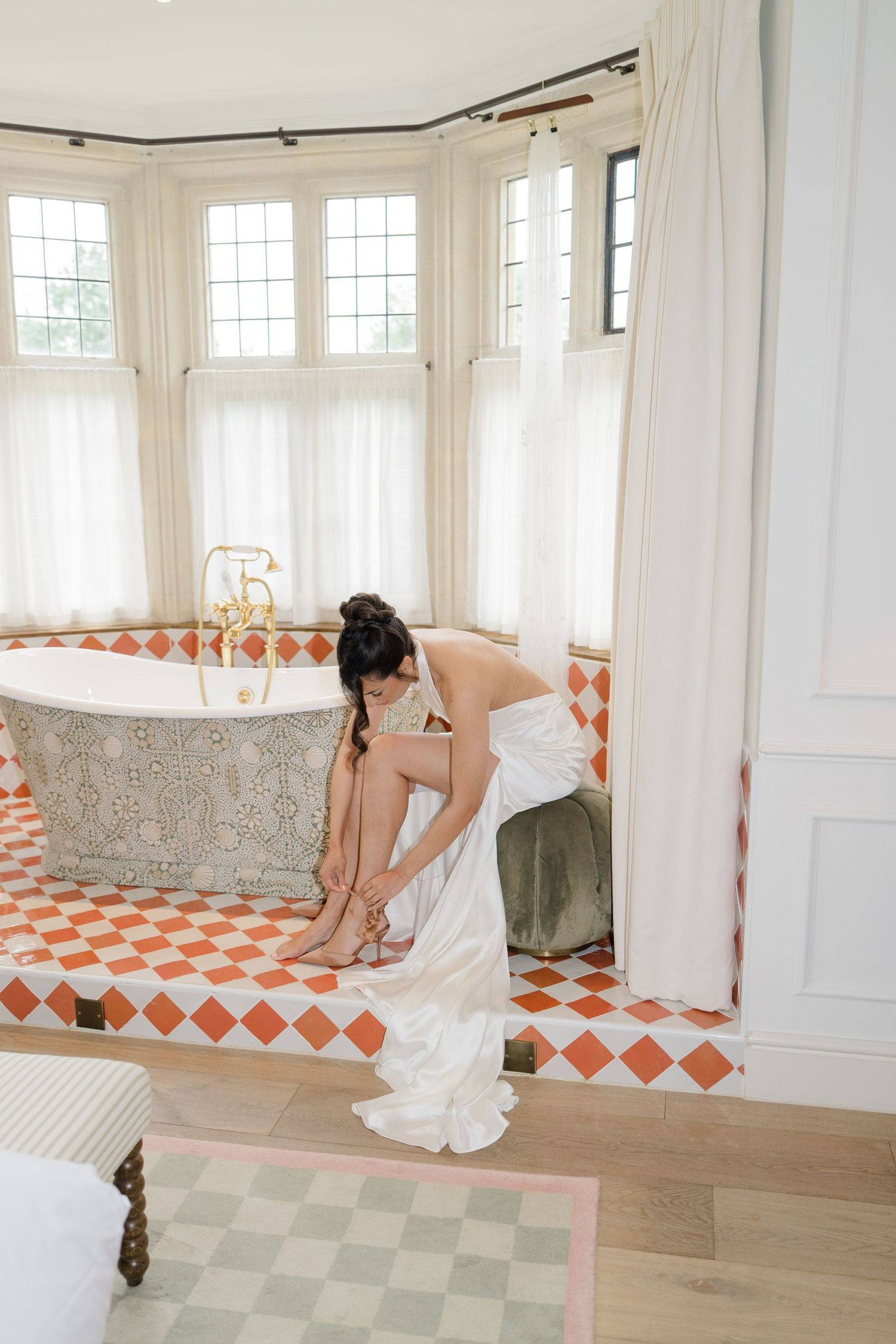 Bride in white wedding gown fastens heels beside decorative clawfoot tub on diamond-patterned tile floor.