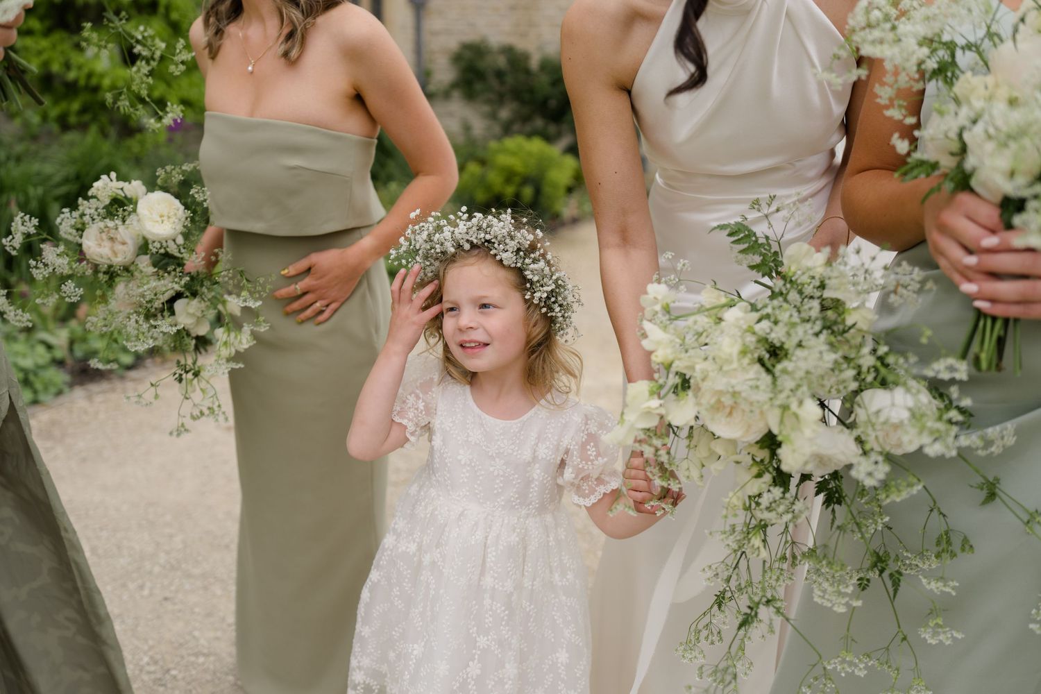 Adorable flower girl in floral crown laughs while holding bridesmaid's hand at outdoor wedding.
