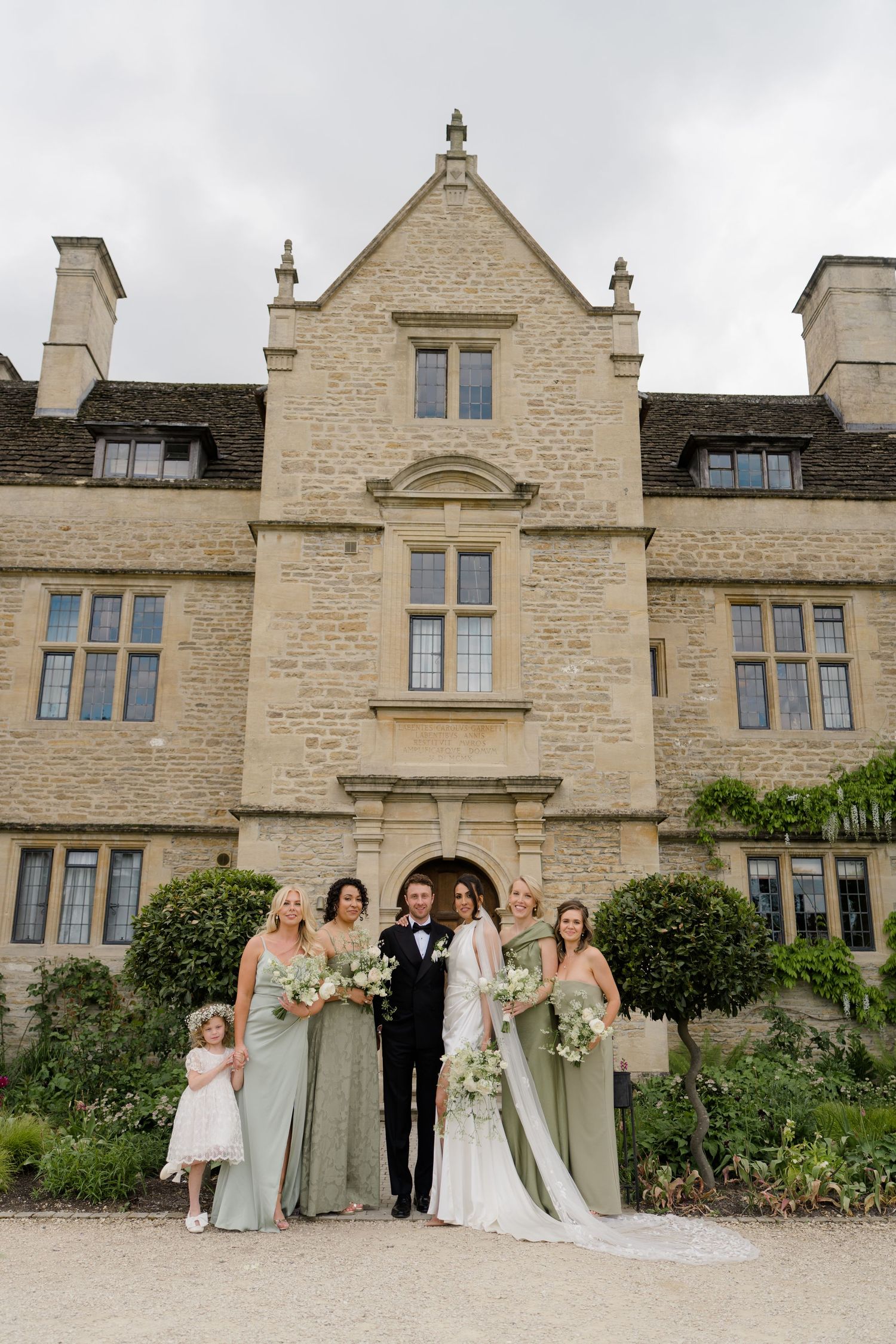 Wedding party poses before grand stone manor house with bridesmaids in sage green gowns holding white bouquets.