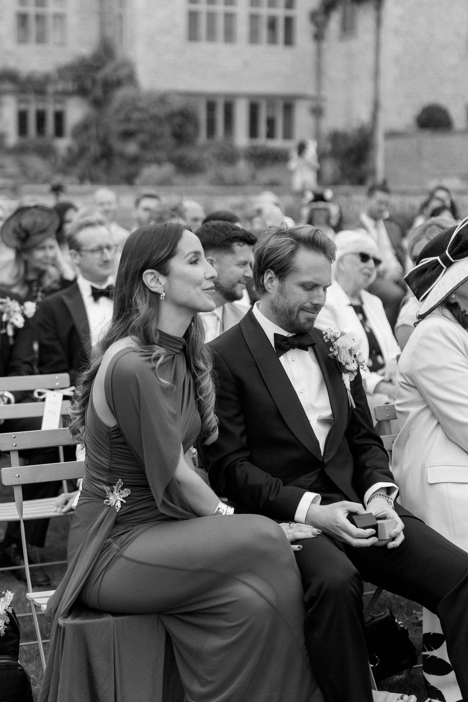 Elegant wedding guests seated outdoors in black and white, woman in gown and man in tuxedo watching ceremony.