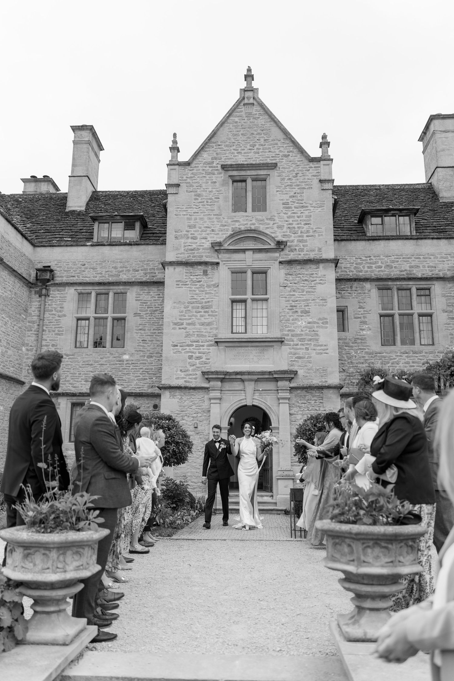 Newlyweds exit through guests outside a grand English stone manor house after their wedding ceremony.