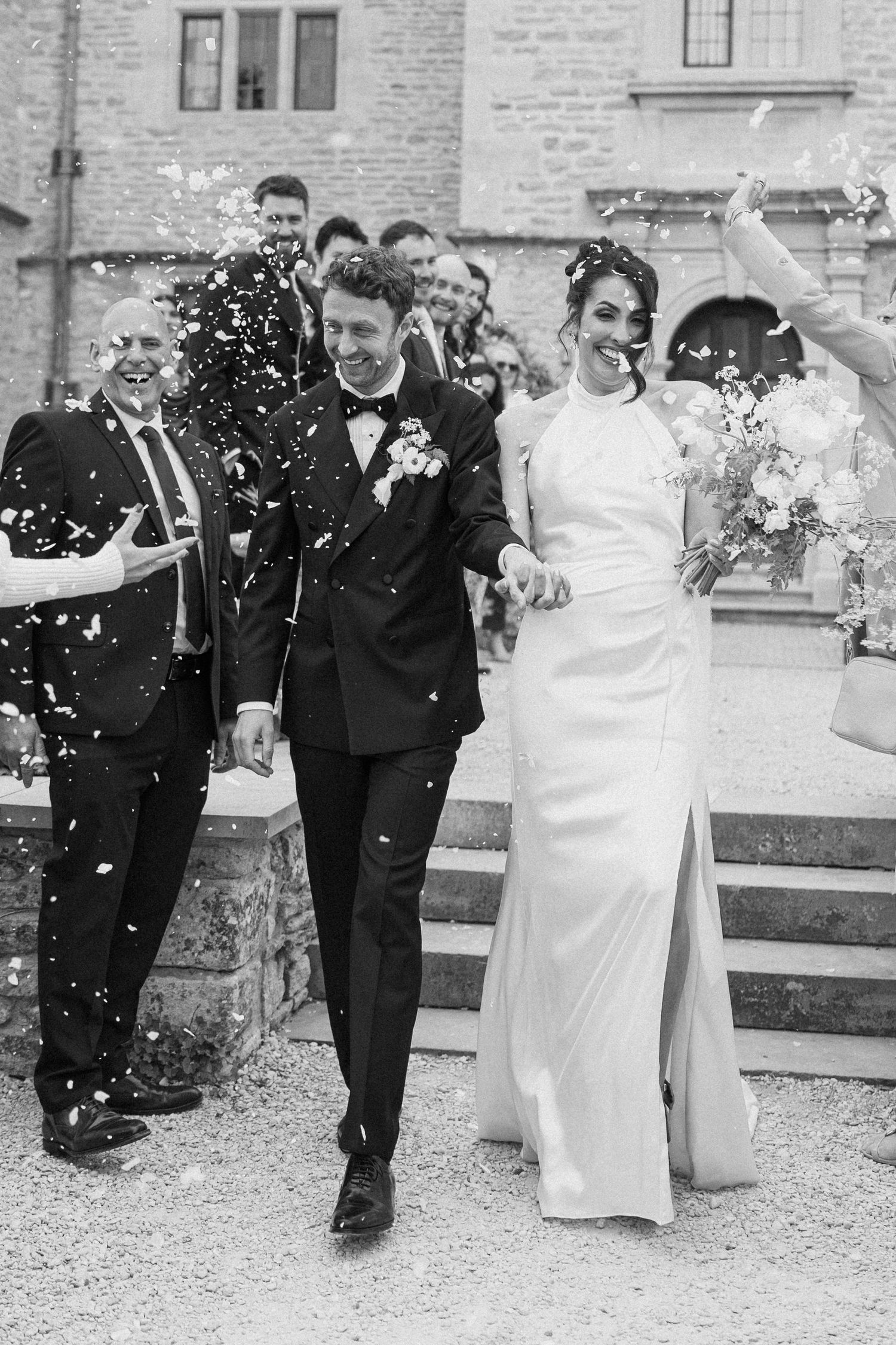 Joyful newlyweds walk through confetti shower outside historic stone venue in black and white photo.