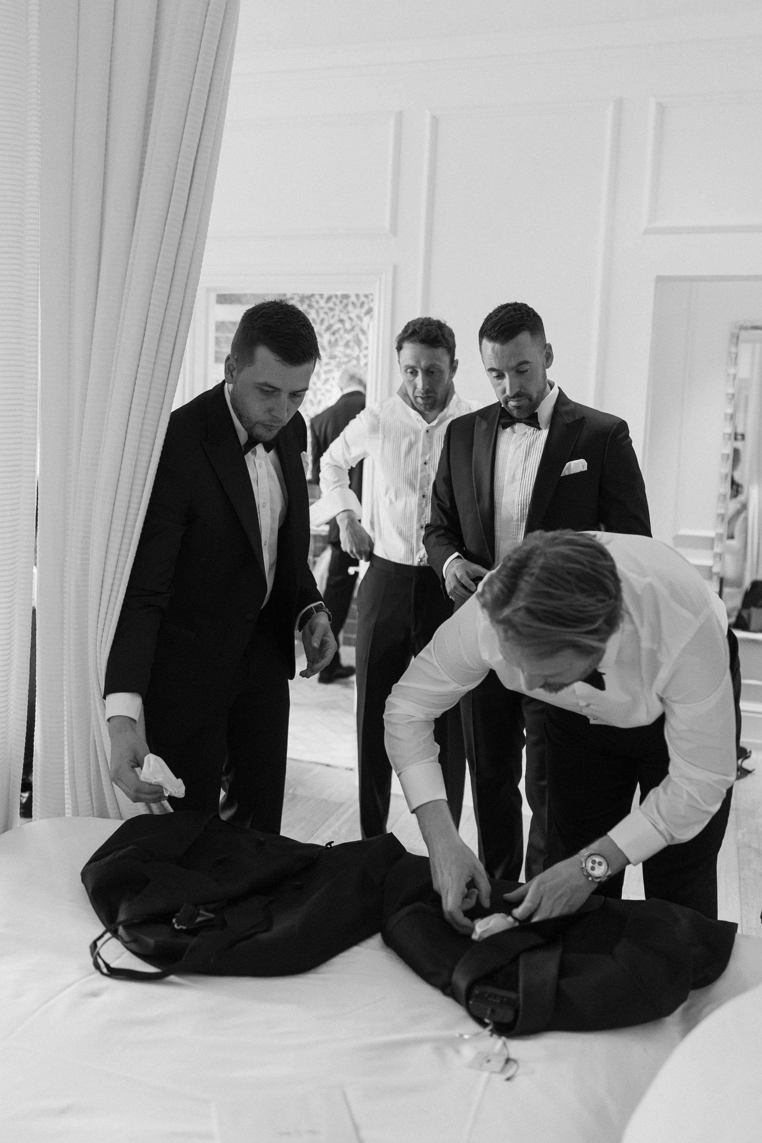Black and white photo of groomsmen in tuxedos preparing suits on a bed before a wedding.