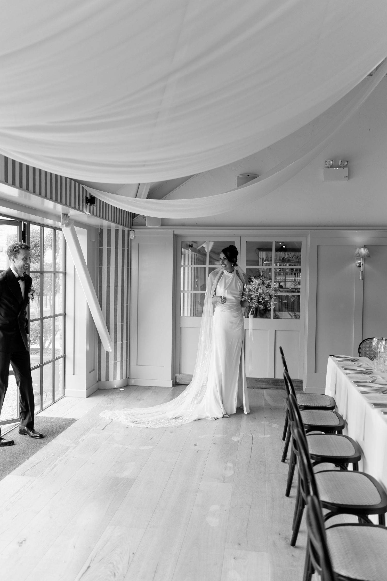 Black and white photo of bride in satin gown standing by French doors in elegantly draped wedding reception room.