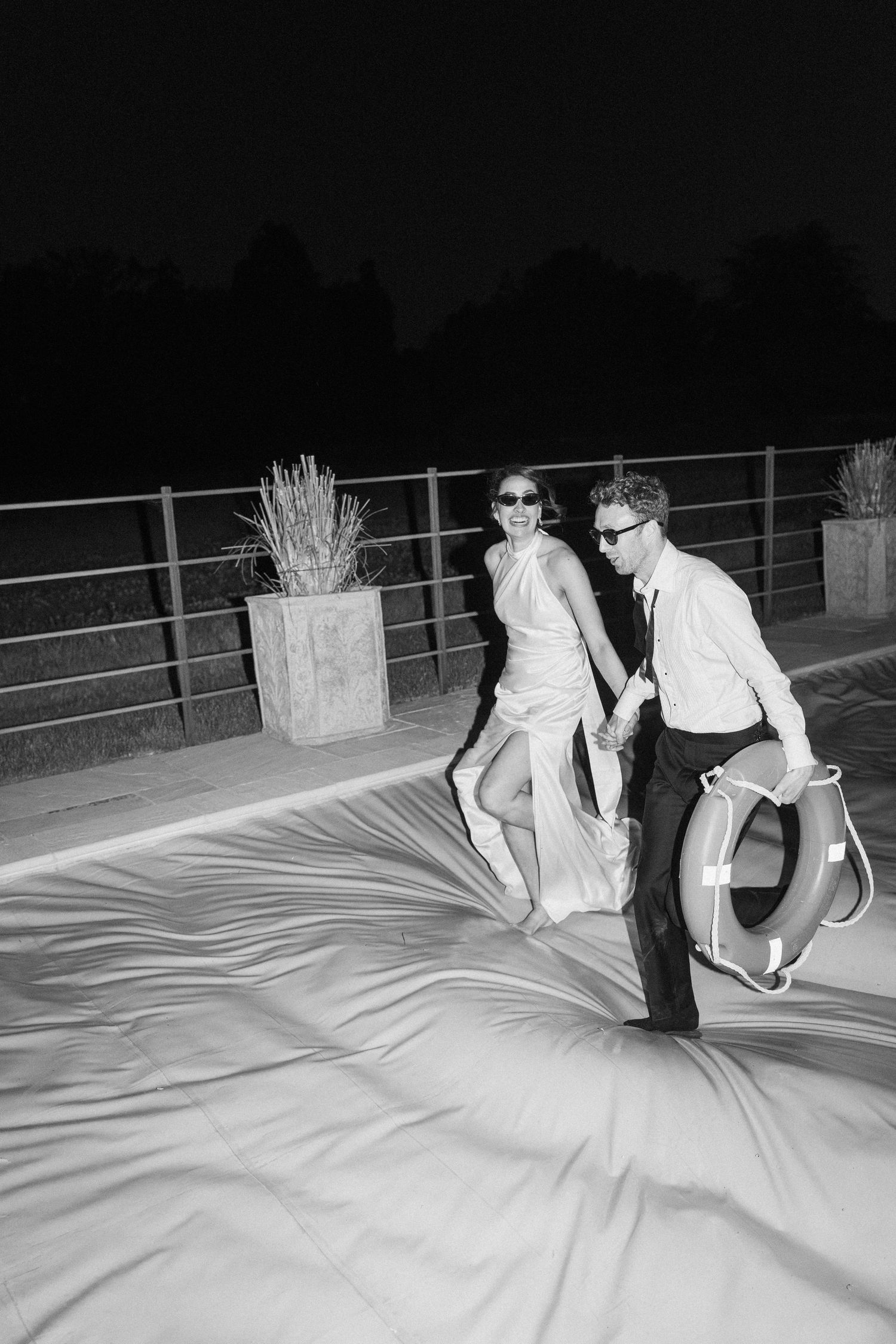 Black and white photo of couple walking on covered pool at night after wedding.