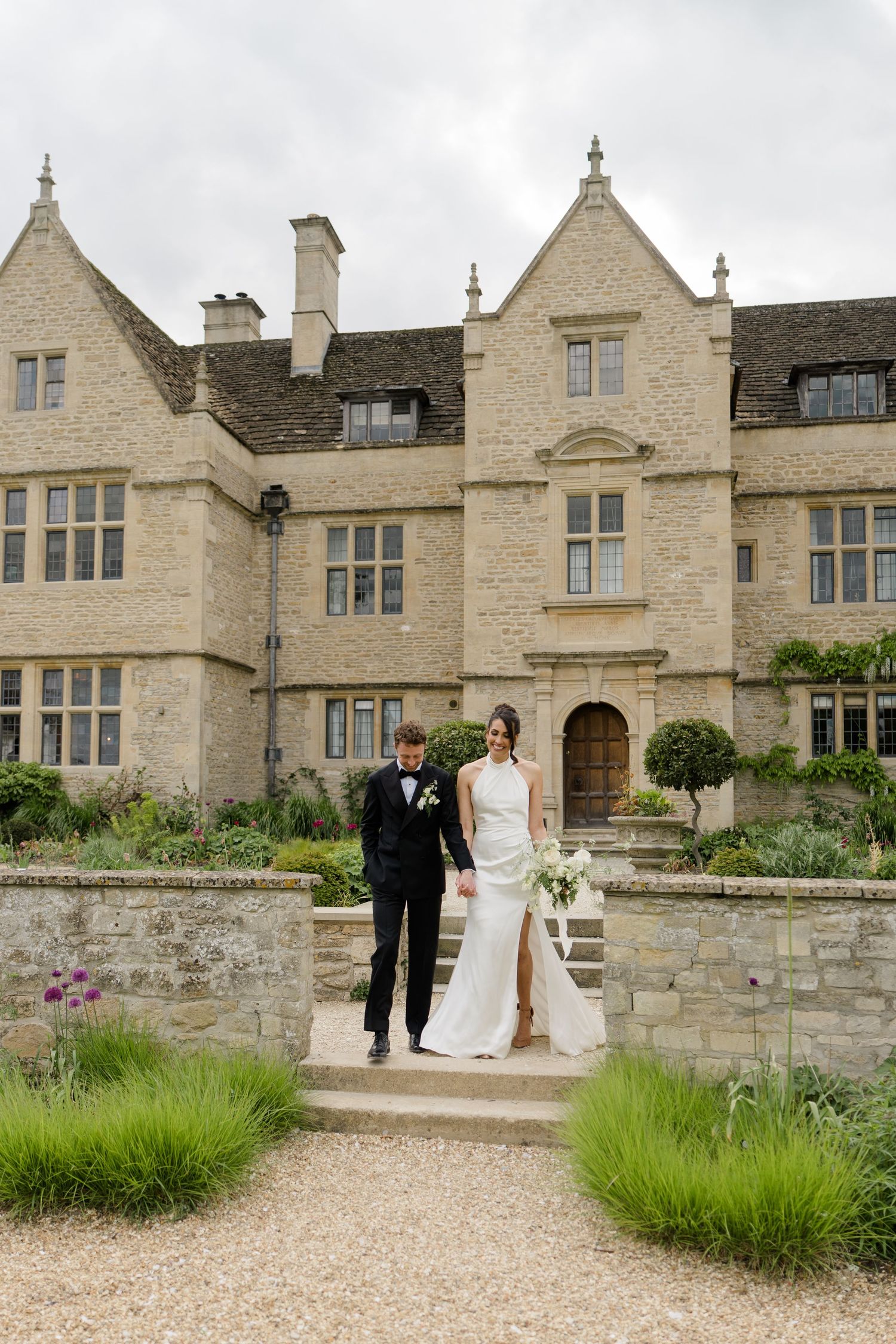 Elegant bride and groom pose on stone steps before a grand Cotswolds manor house on their wedding day.