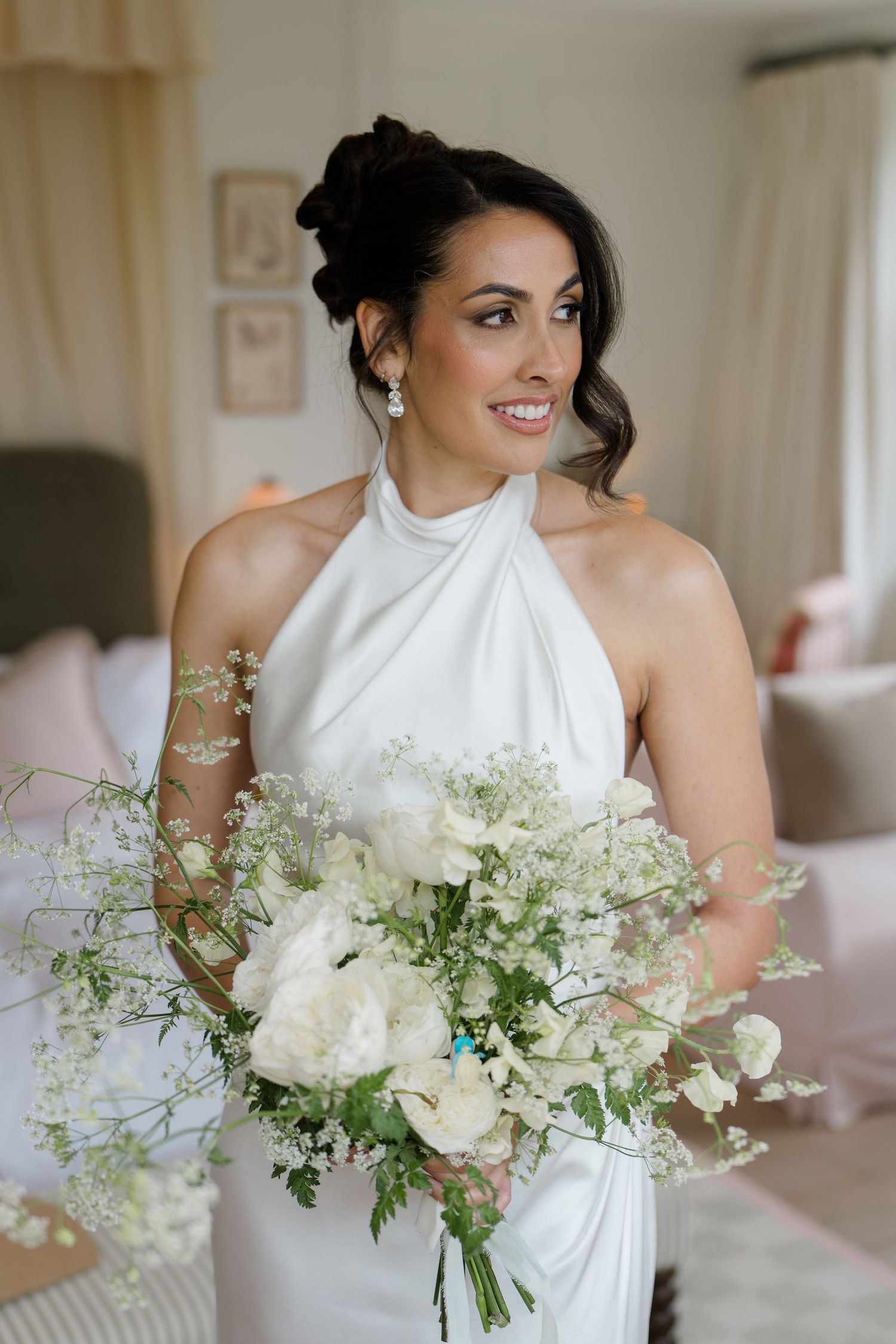 Bride in elegant white halter neck dress holds a lush white and green bouquet in a bright room.