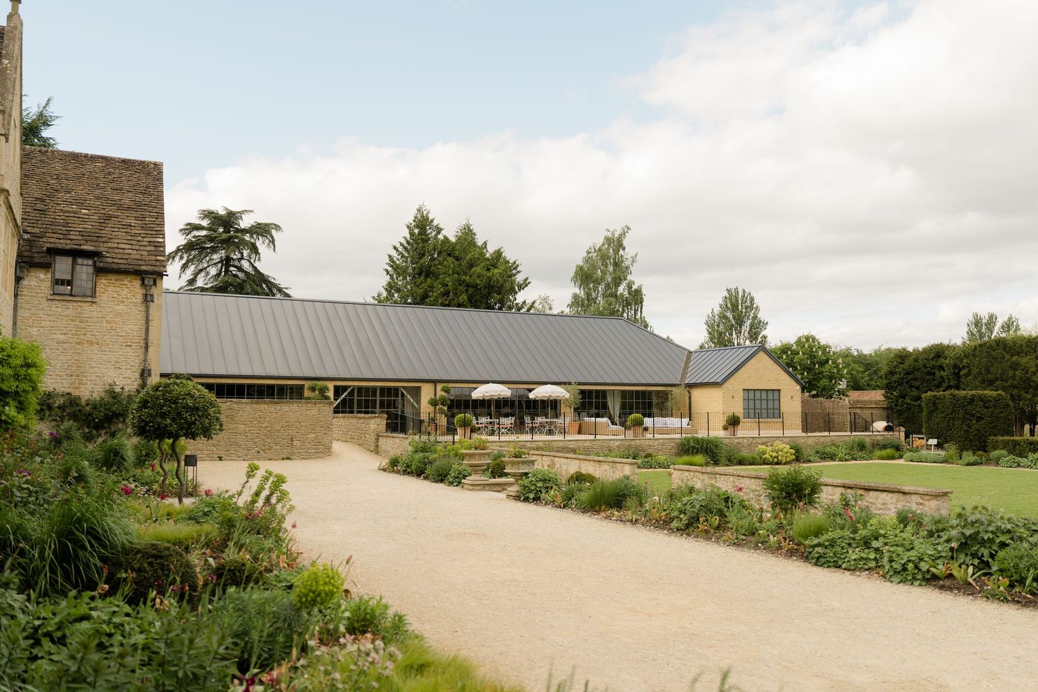 Elegant Cotswold stone venue with manicured gardens and modern barn extension under a cloudy sky.