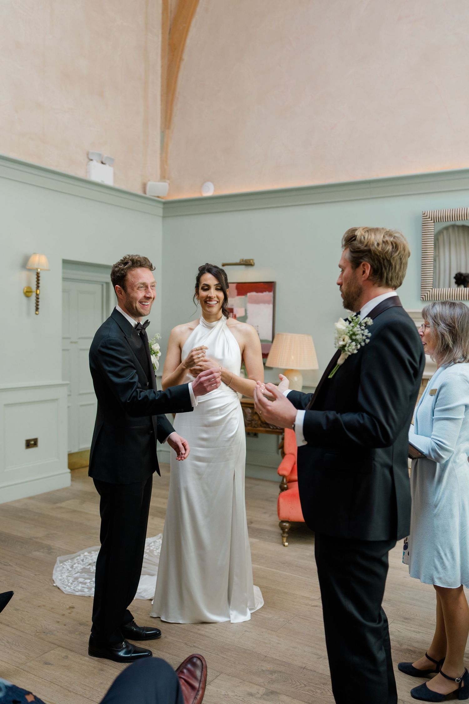 Bride and groom laughing with best man during wedding ring exchange ceremony indoors.