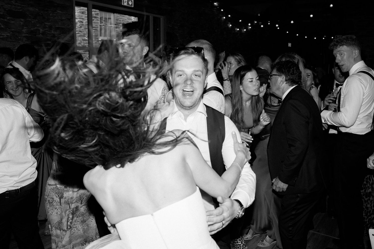 Couples dancing at a wedding reception under string lights in a rustic venue.