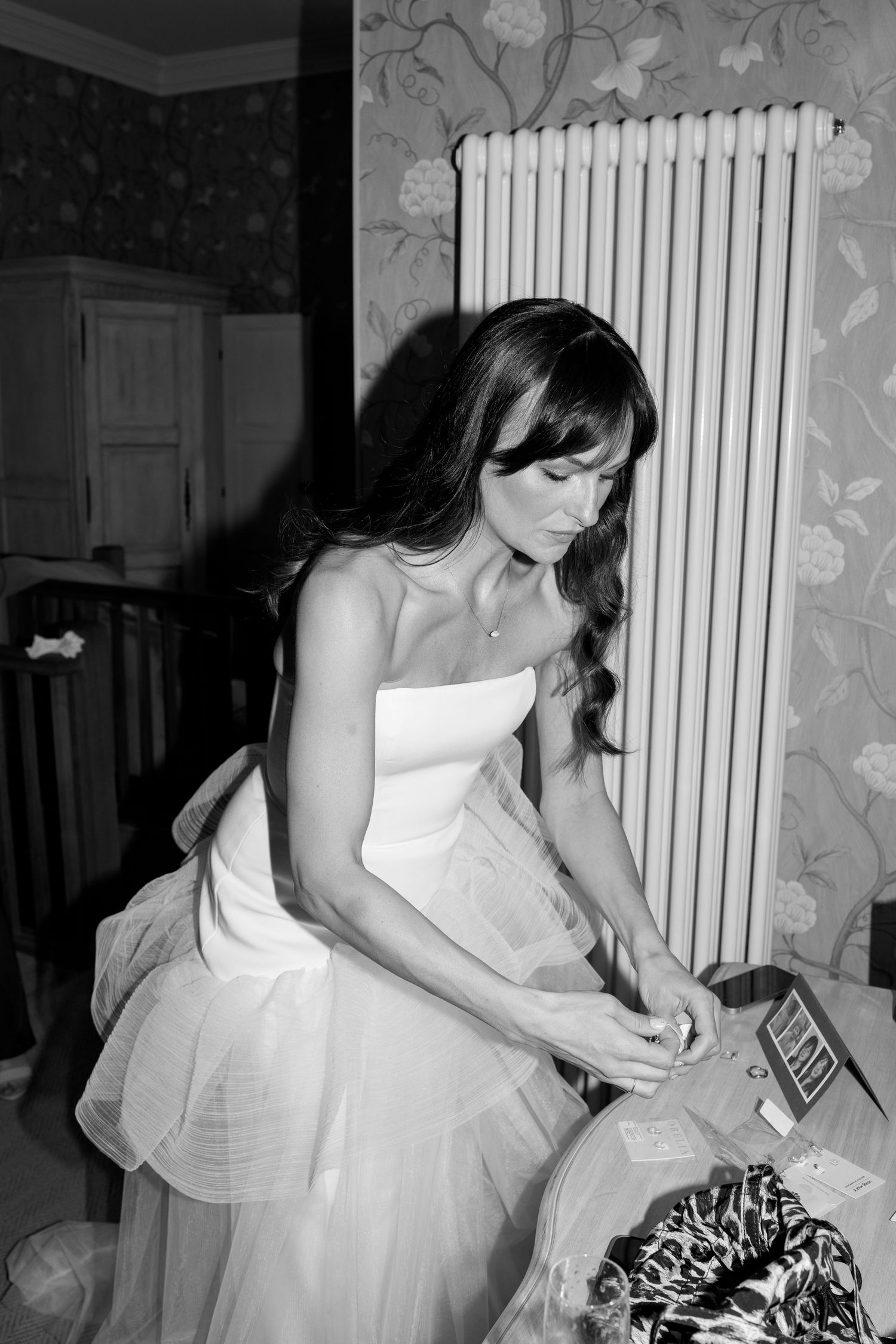 A bride in a strapless wedding dress sits by a radiator during wedding preparations.