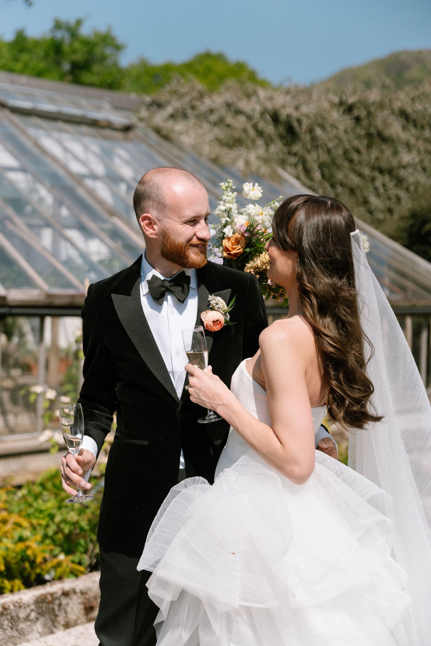 A joyful wedding couple laughs together while holding champagne glasses in a garden setting.