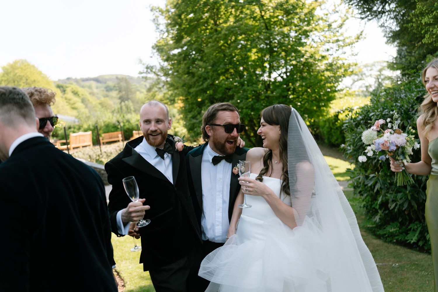 Wedding party members walk together through a garden path on a sunny day.