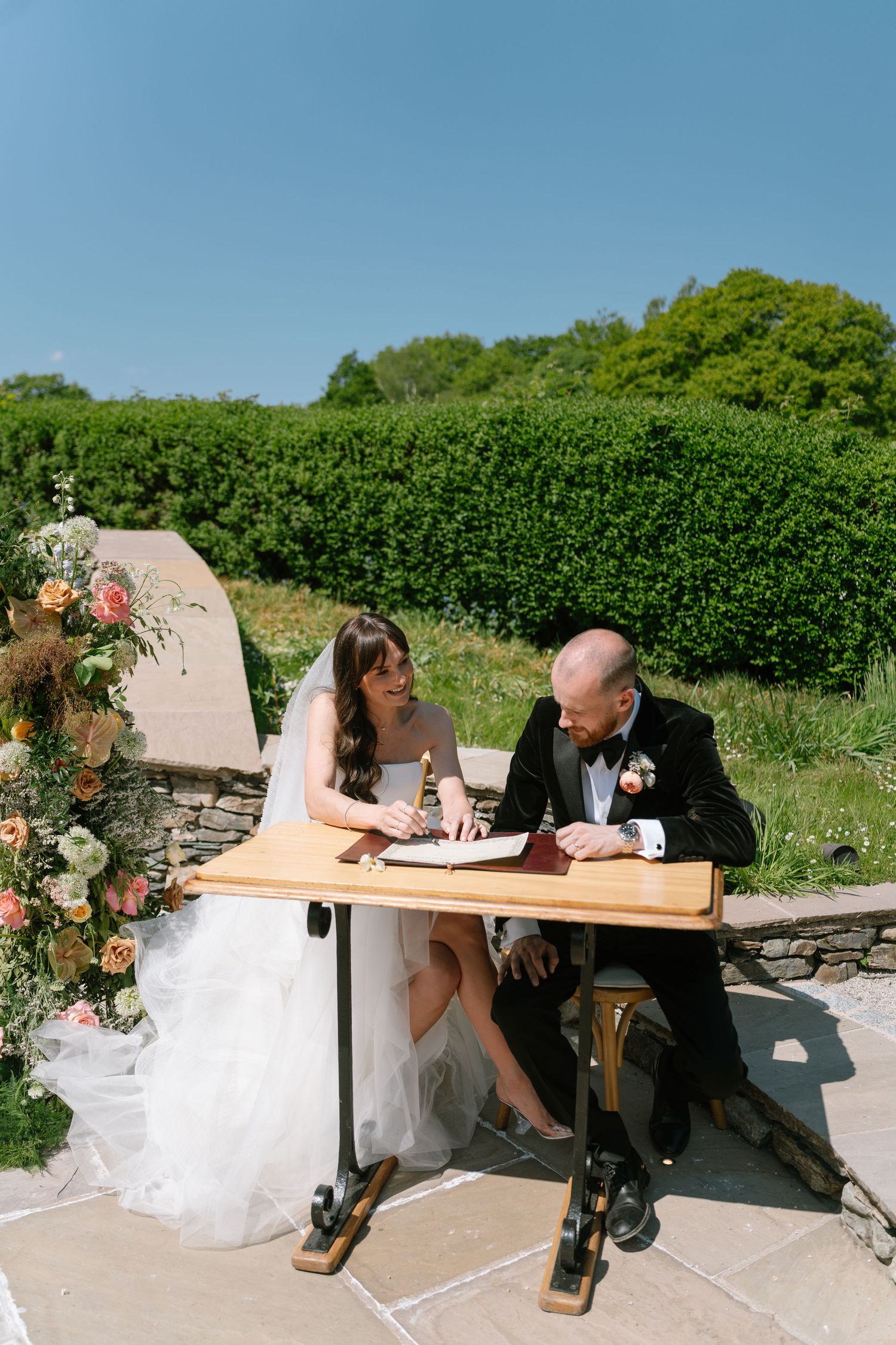 Newlyweds sign their marriage certificate at an outdoor table on a sunny day.