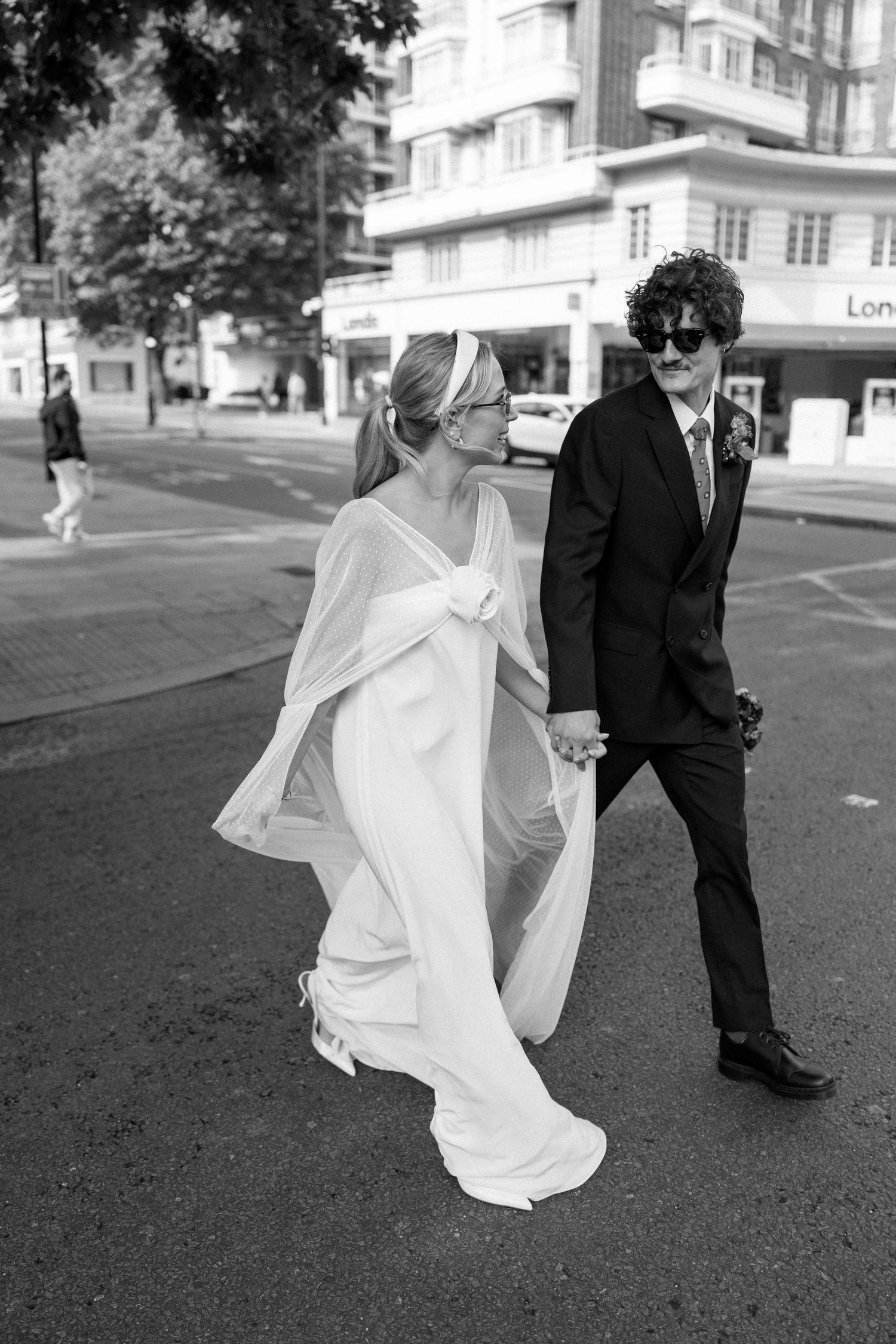 A flowing white wedding dress billows in the wind as a couple walks across a paved outdoor area.