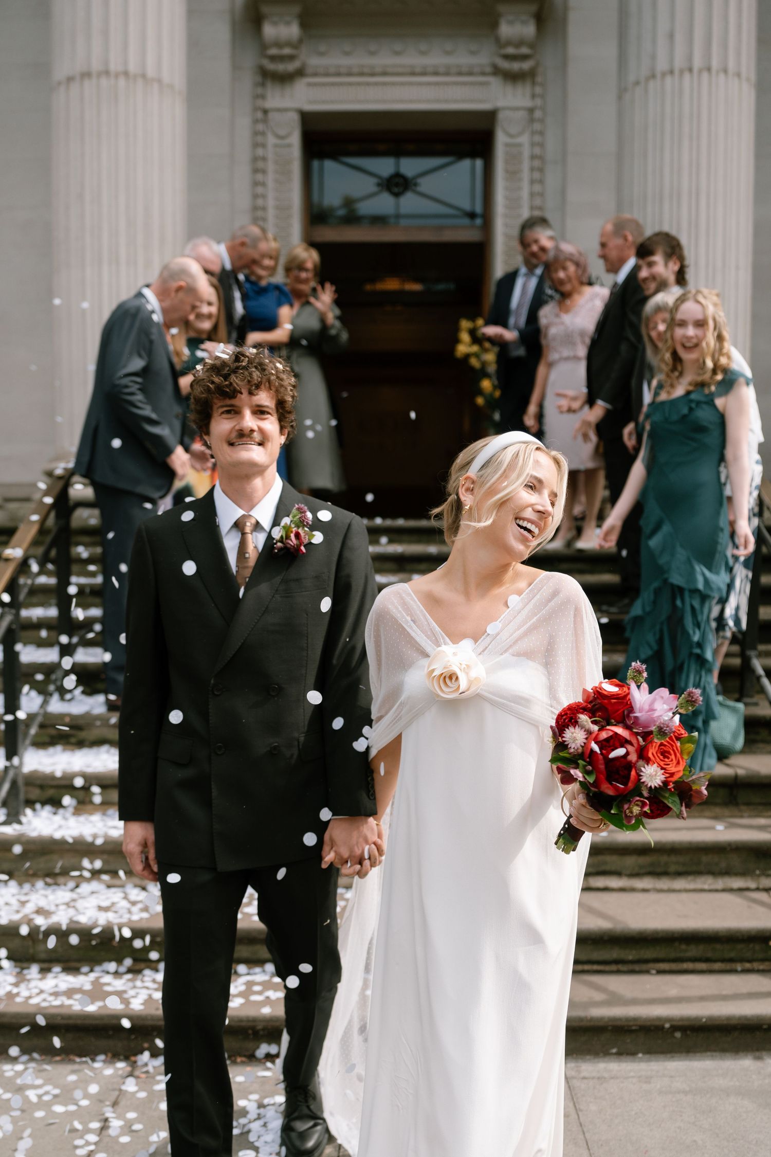 Newlyweds share joyful moment amid confetti shower outside historic venue.