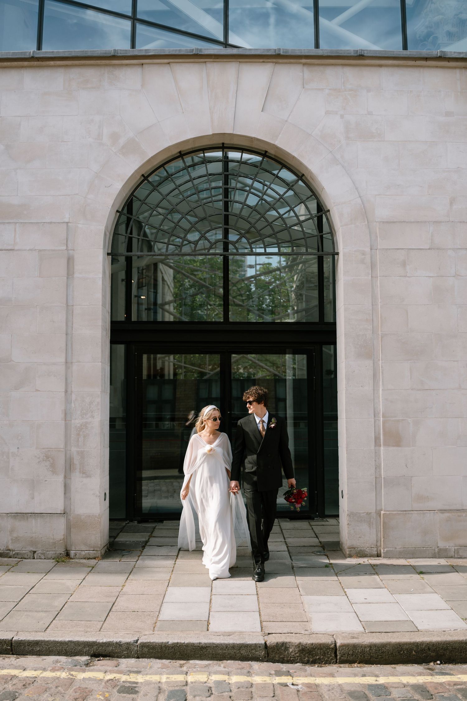 A newlywed couple exits through an elegant arched glass doorway of a stone building.
