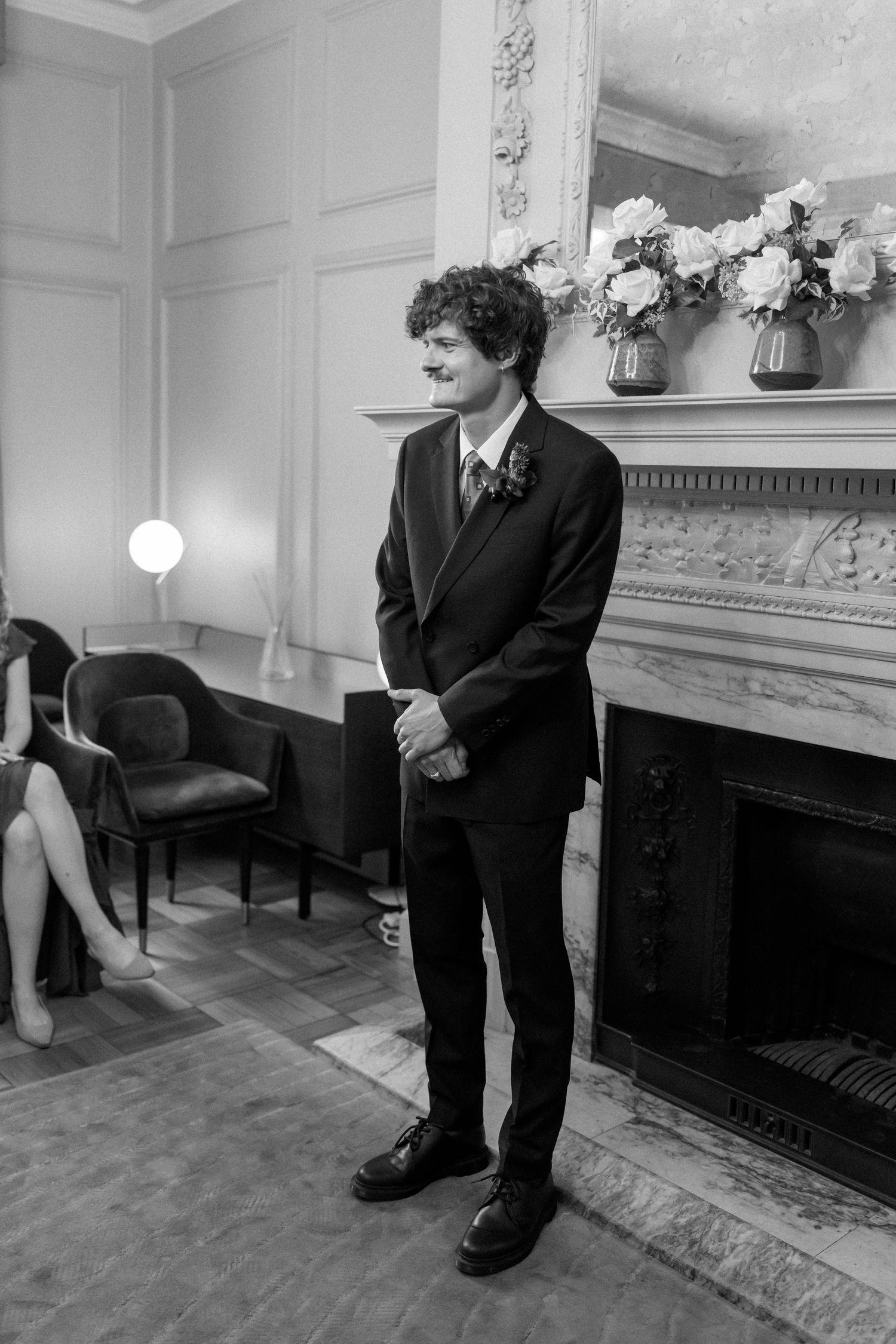 A person in formal attire standing by a fireplace with floral arrangements in a classic room.