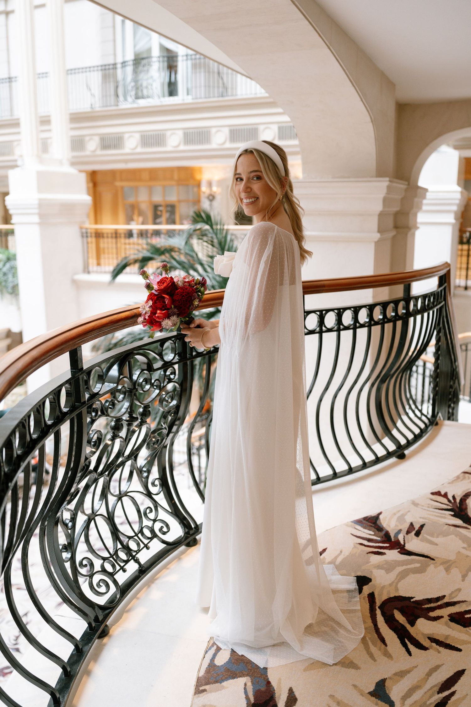 A bride in a flowing white gown poses elegantly along an ornate curved staircase with black iron railings.
