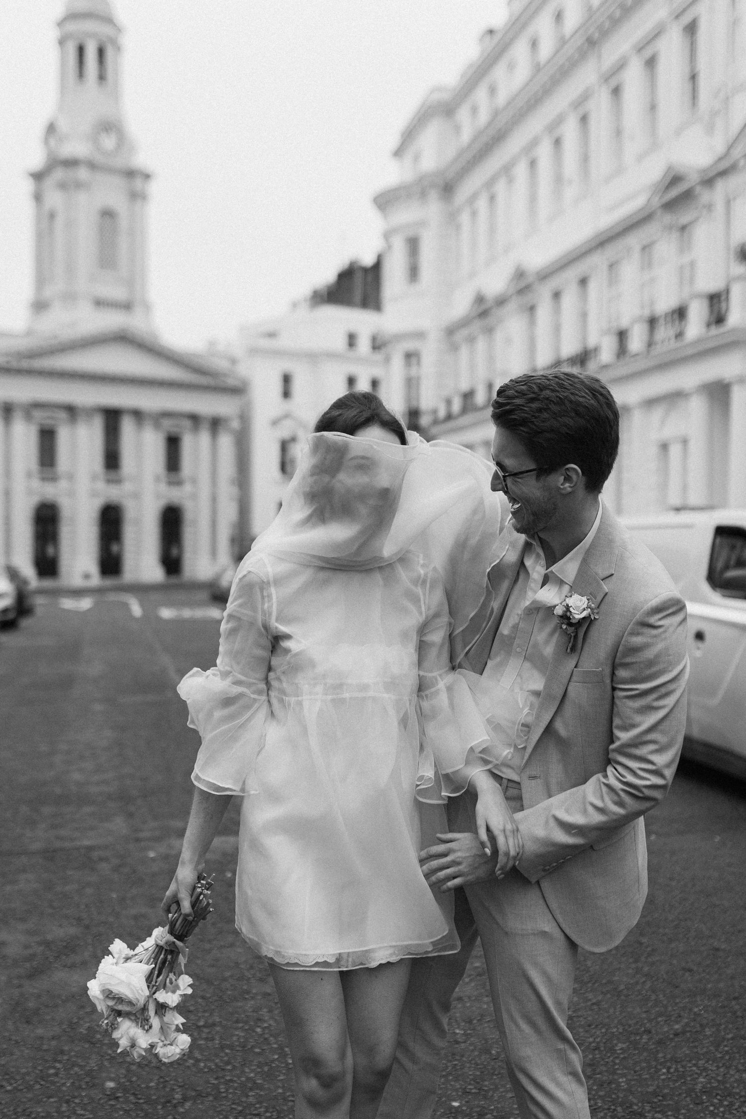 Black and white photo of a bride in short dress and groom in suit walking together on a city street after their wedding.