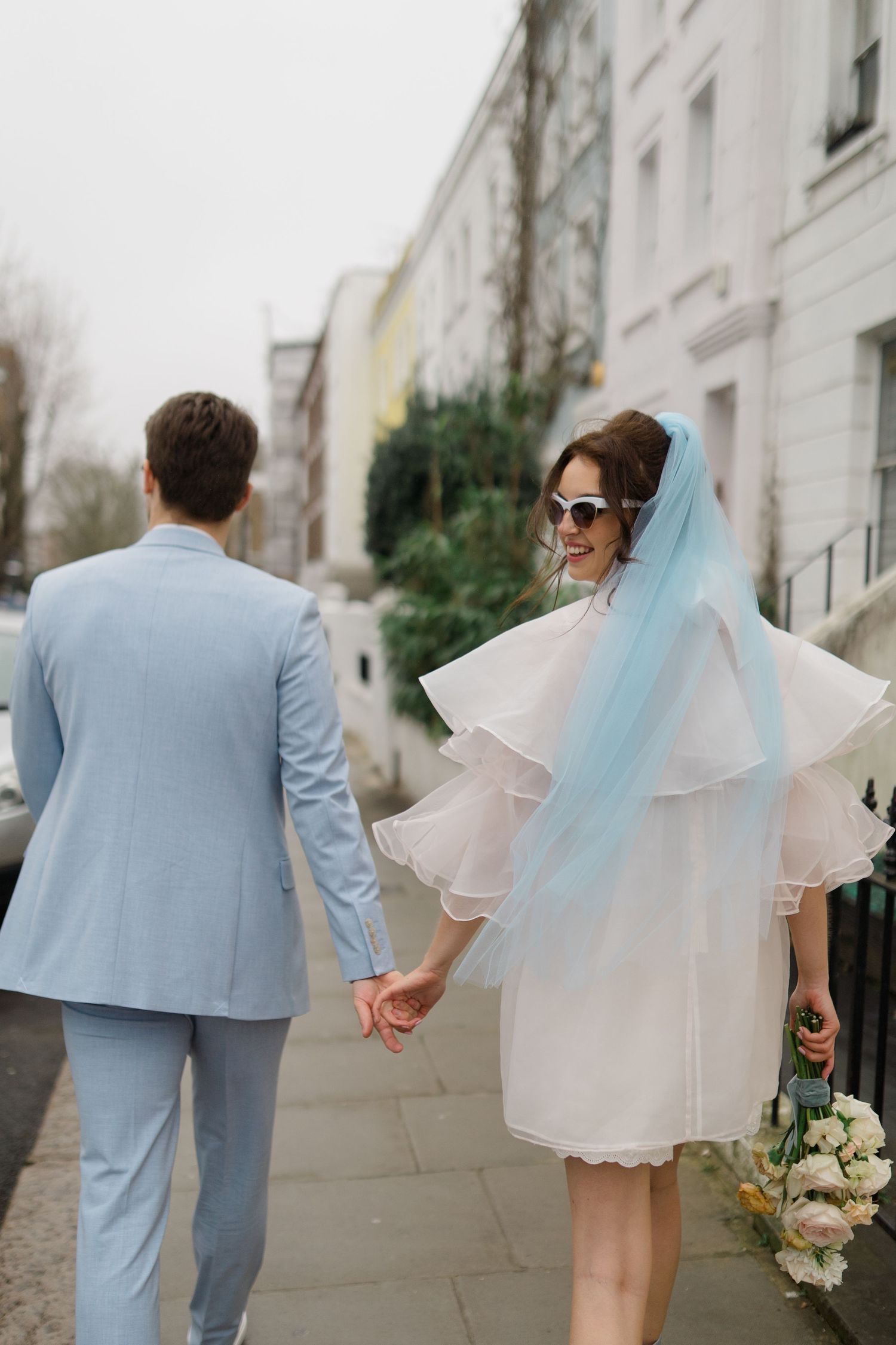 Bride in short white dress with blue veil and groom in light blue suit walking hand in hand on a London street.