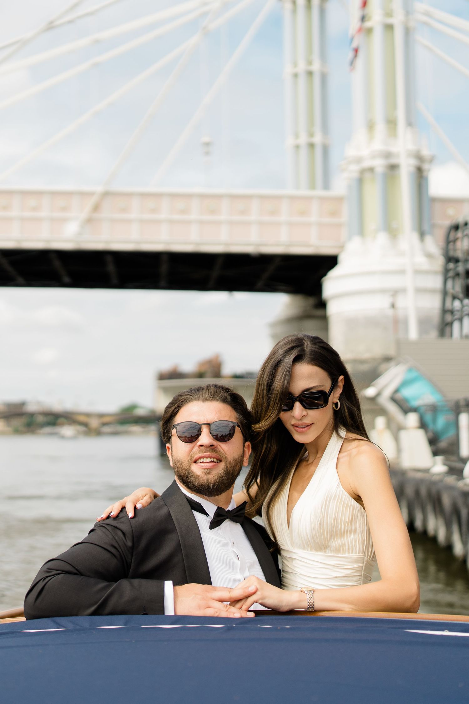Elegant couple in formal attire and sunglasses pose on a luxury boat with a bridge backdrop on the Thames.