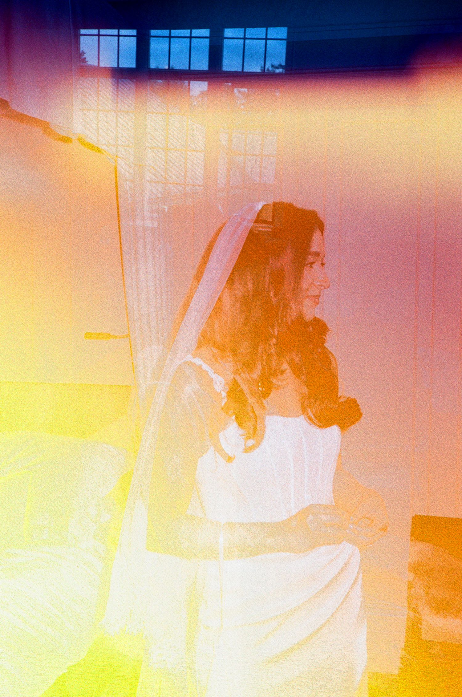A bride in a white dress and veil stands in a warmly lit room, captured with a dreamy film light leak effect.