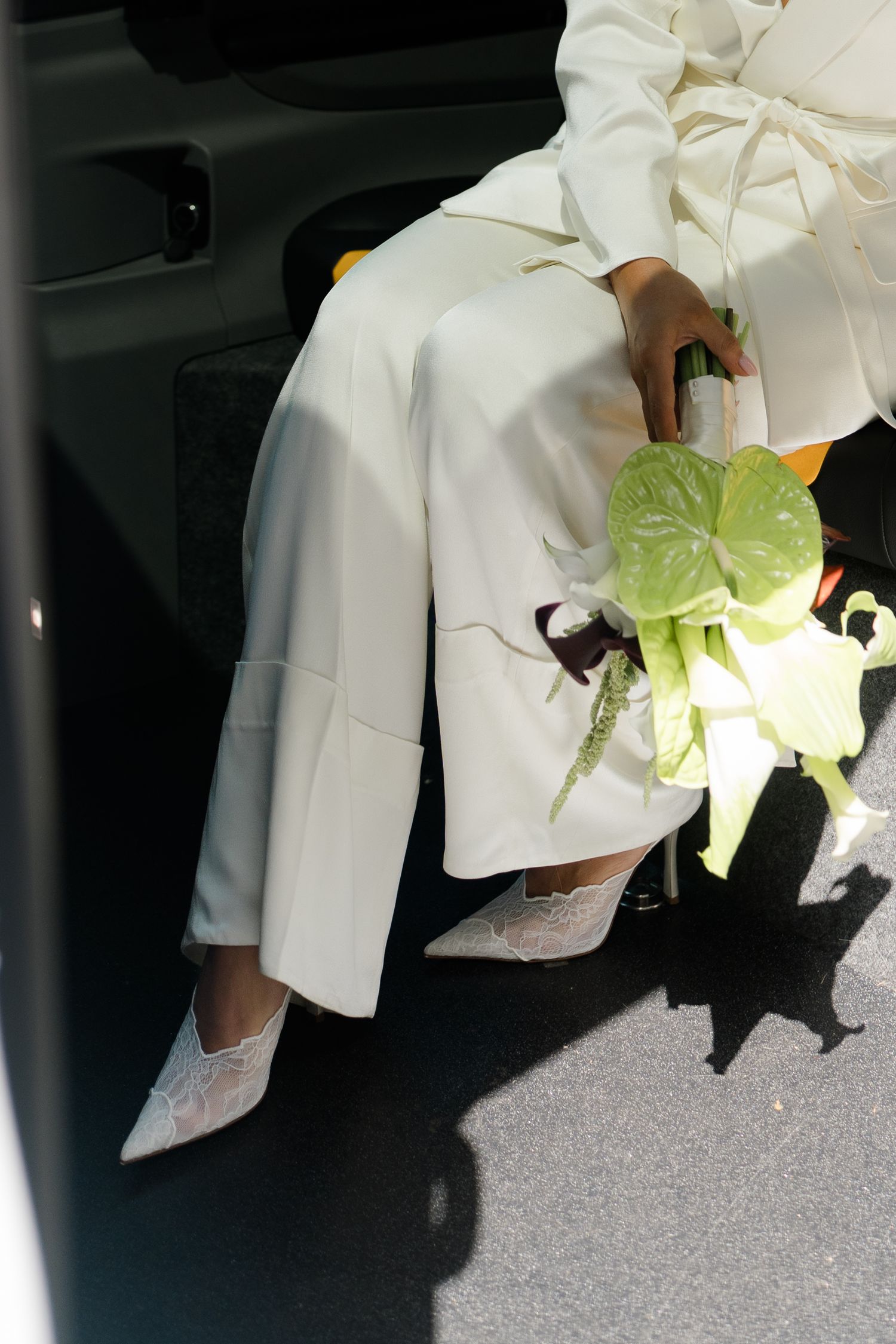 Bride in white pantsuit holding green anthurium bouquet, sitting in car doorway with lace heels.
