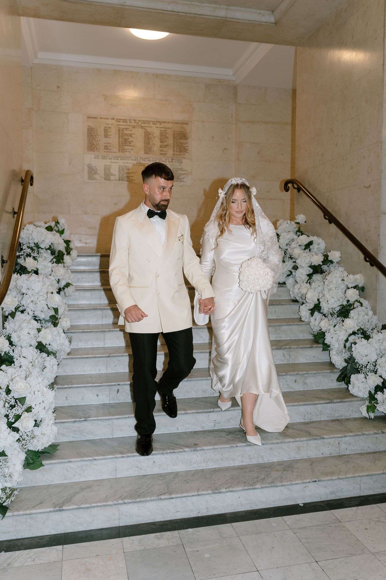 Wedding ceremony participants walking down ornate staircase adorned with white floral decorations.
