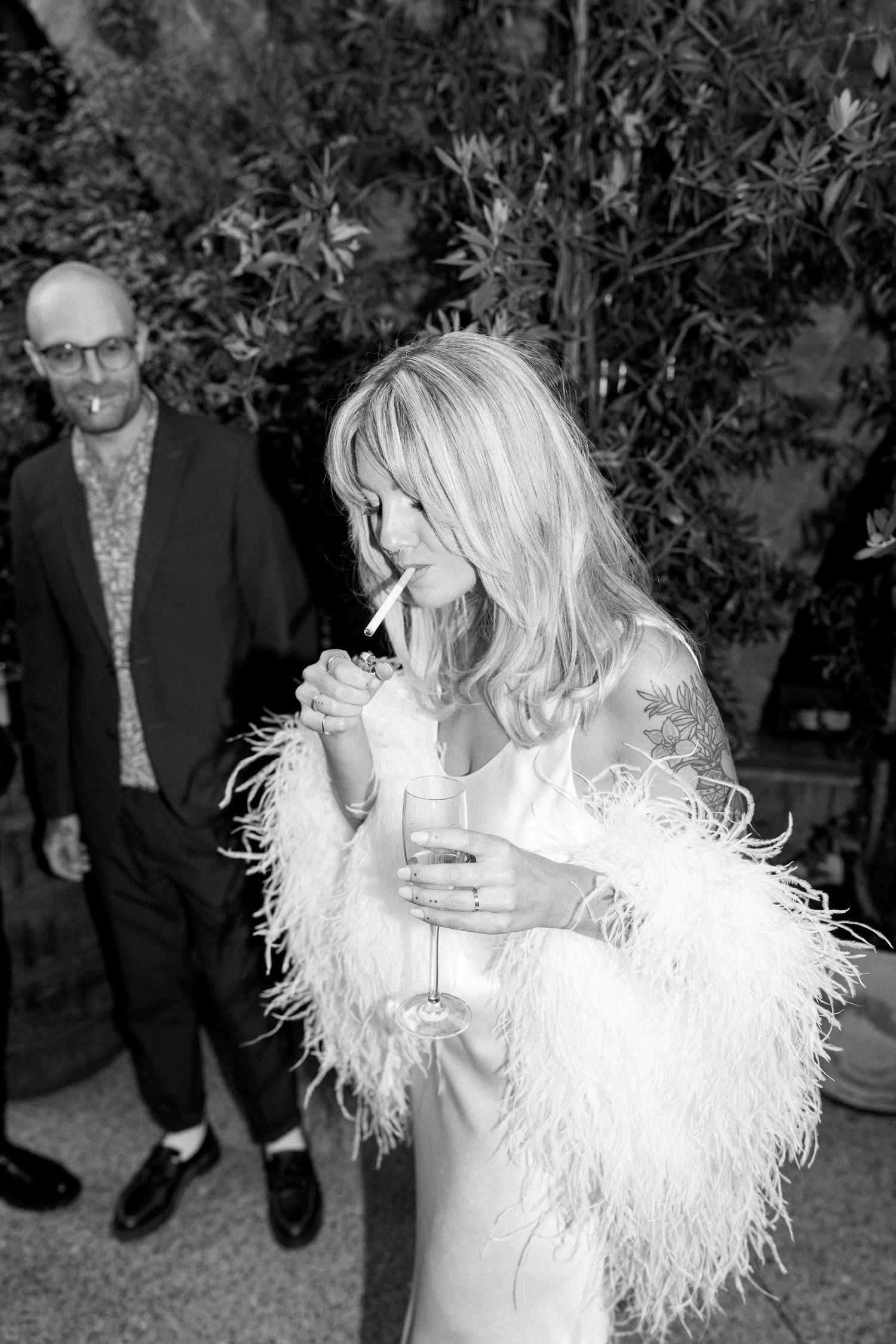 Black and white photo of someone in a white feathered dress holding a champagne glass at an outdoor event.