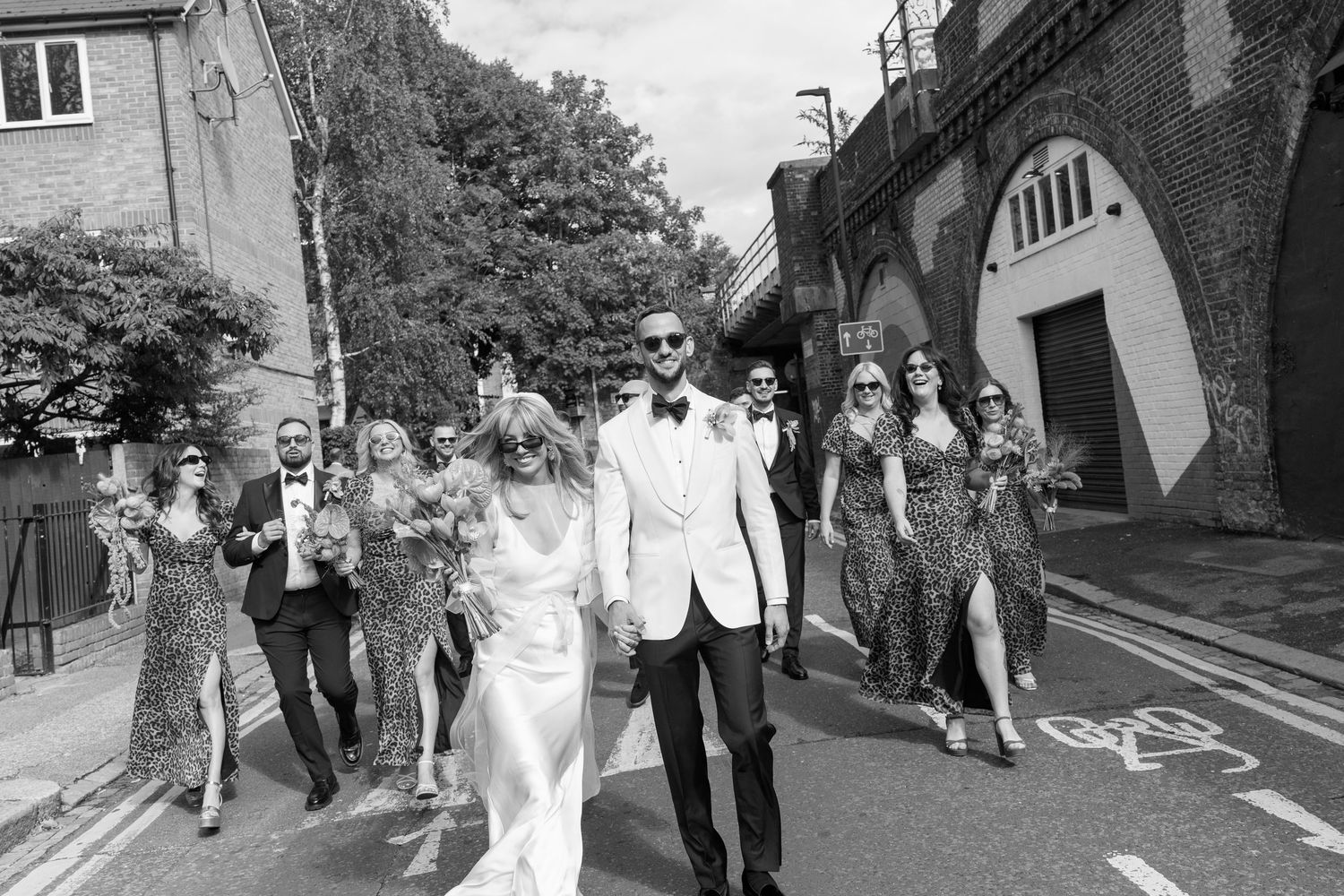 Wedding party and guests walk down a street in formal attire.