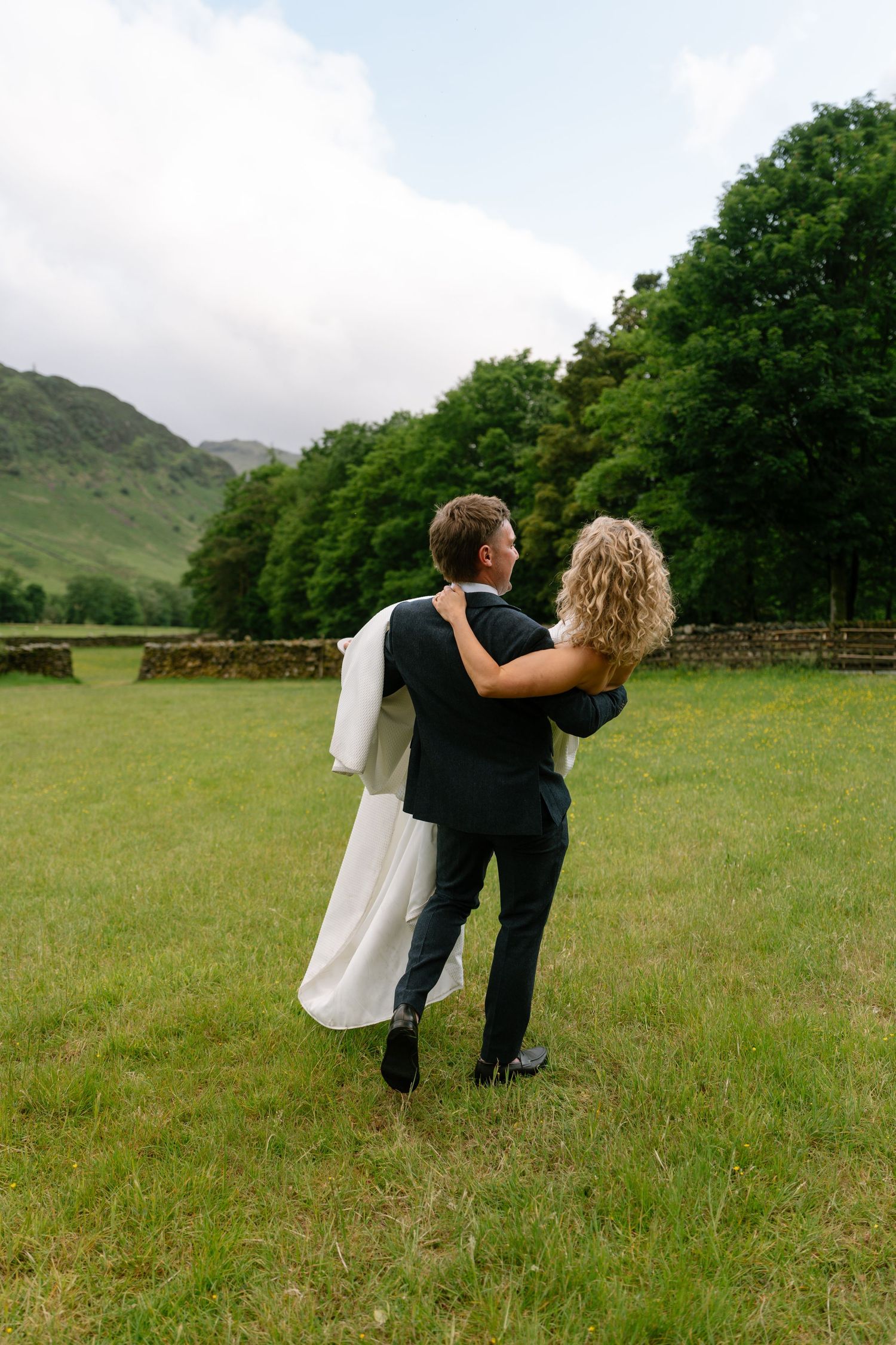 Couple walks through a green meadow with mountains in the background.