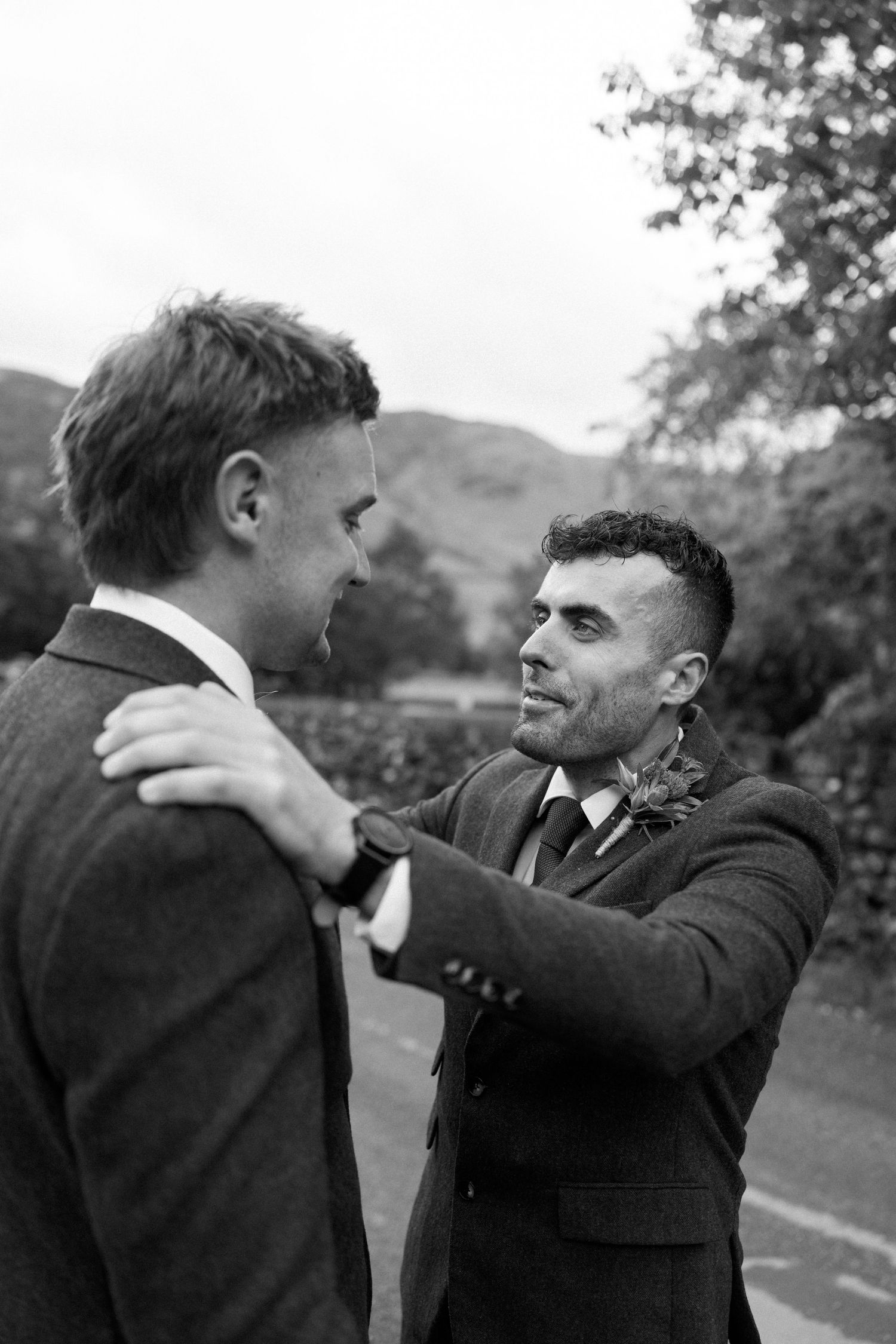 Two men in suits adjusting each other's formal attire outdoors with mountains in the background.