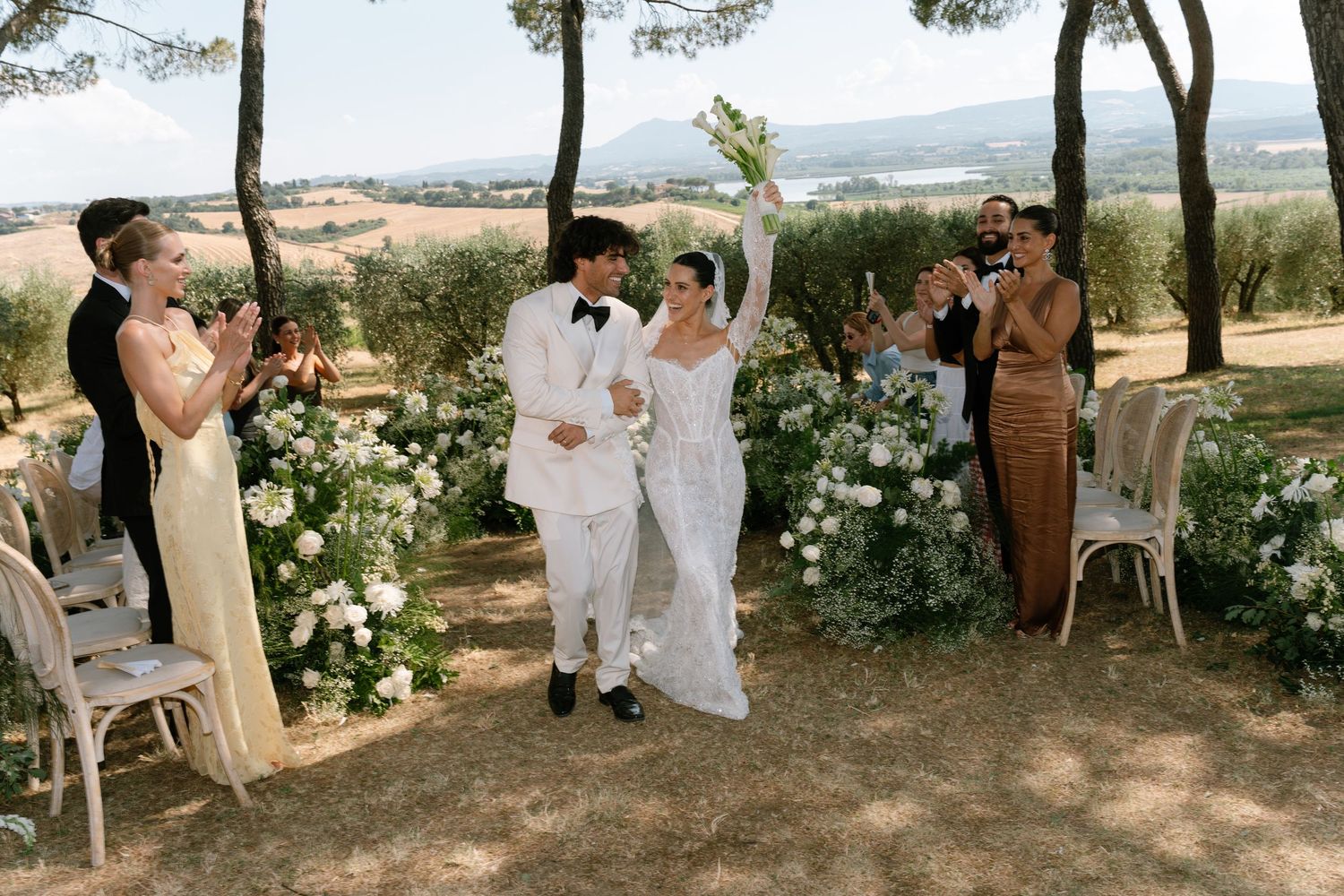 Outdoor wedding celebration with guests gathered under trees and white floral decorations.
