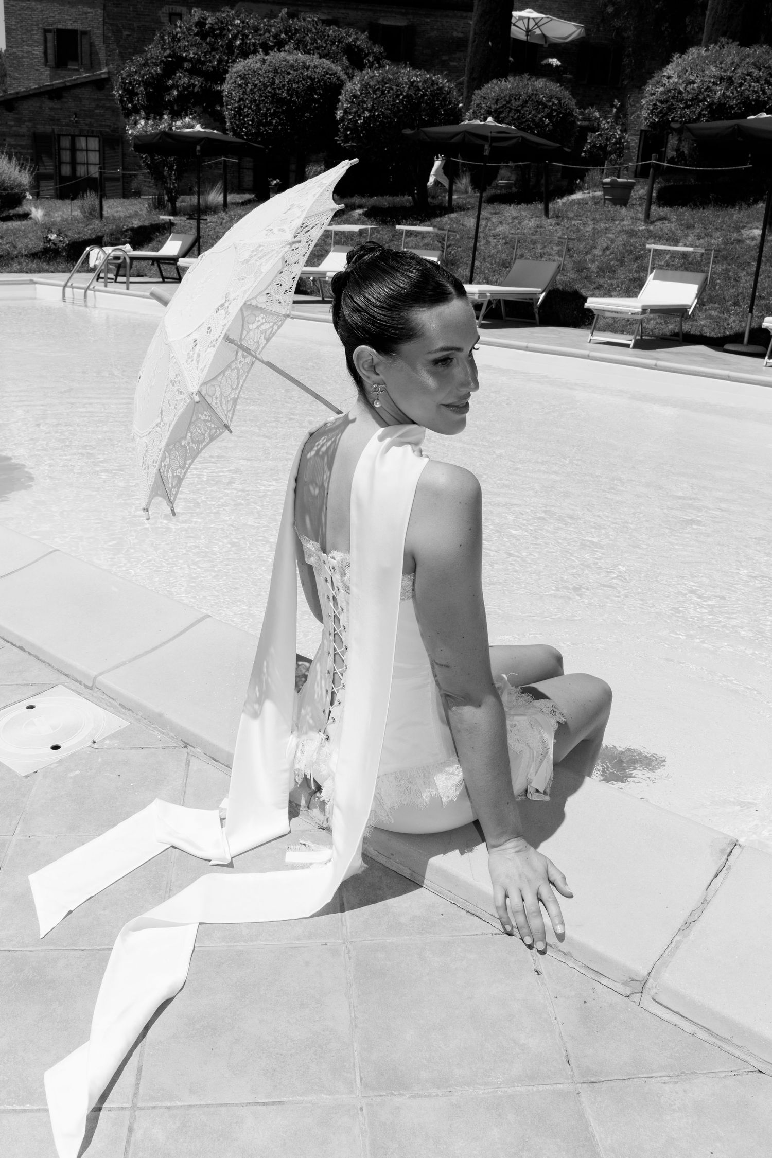 Person in white dress holding parasol sitting by poolside in black and white photograph.