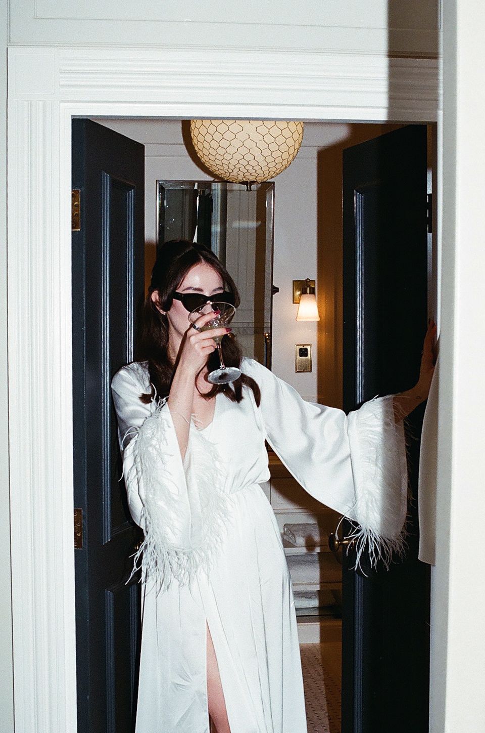 Person in white feathered robe and sunglasses poses playfully in three different stances in a hotel hallway.