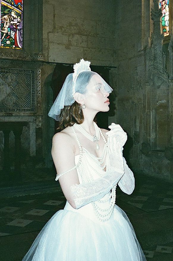 A person in a white wedding dress and pearls poses dramatically in a gothic cathedral interior.