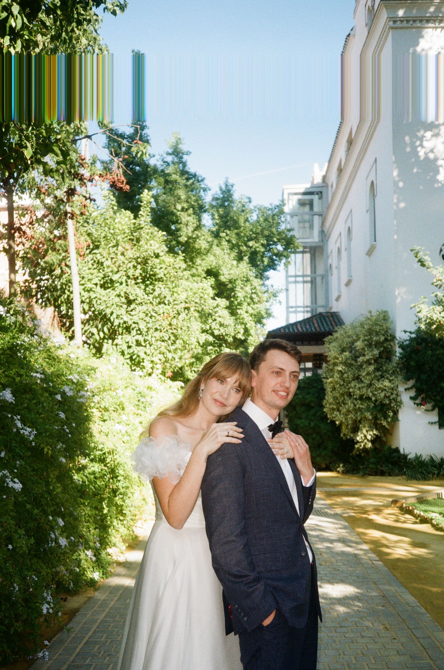 A couple poses together outdoors on a sunny walkway with white buildings and greenery in the background.