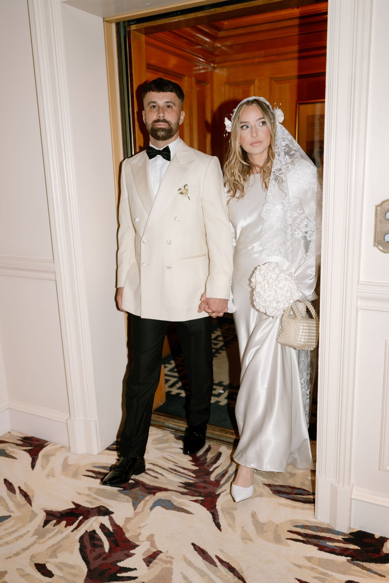 A newlywed couple stands in a doorway wearing white formal attire.