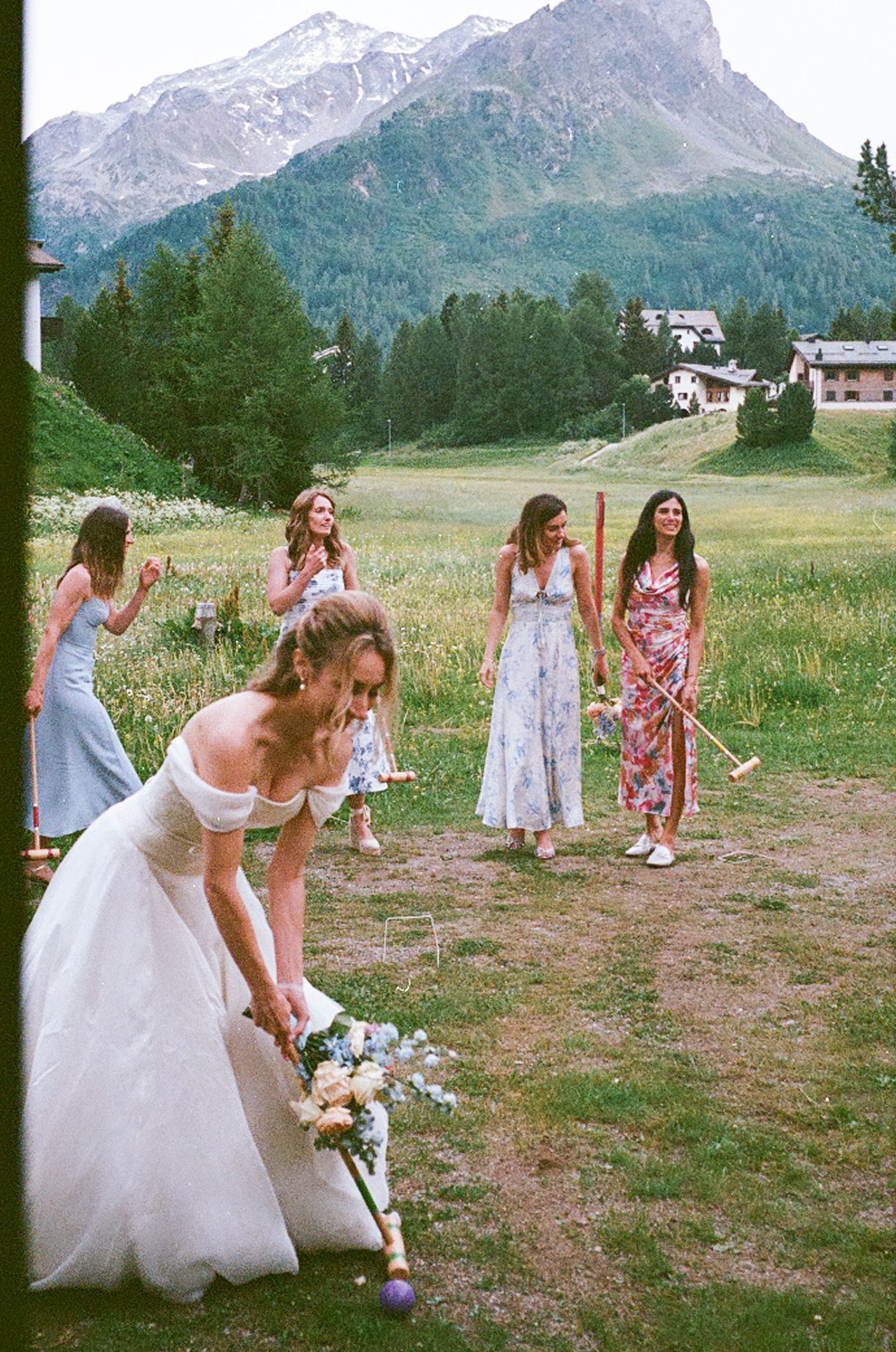 A wedding celebration takes place in a scenic mountain meadow with snow-capped peaks and alpine forests in the background.