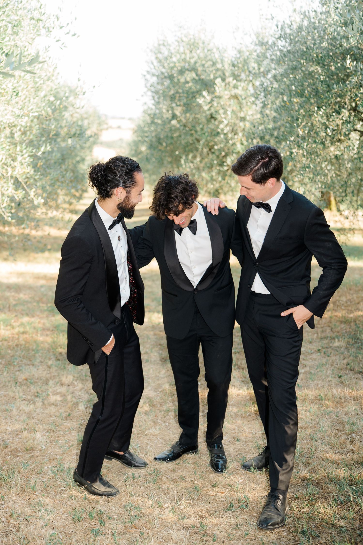Three groomsmen in black tuxedos and bow ties share a candid moment together in an olive grove.