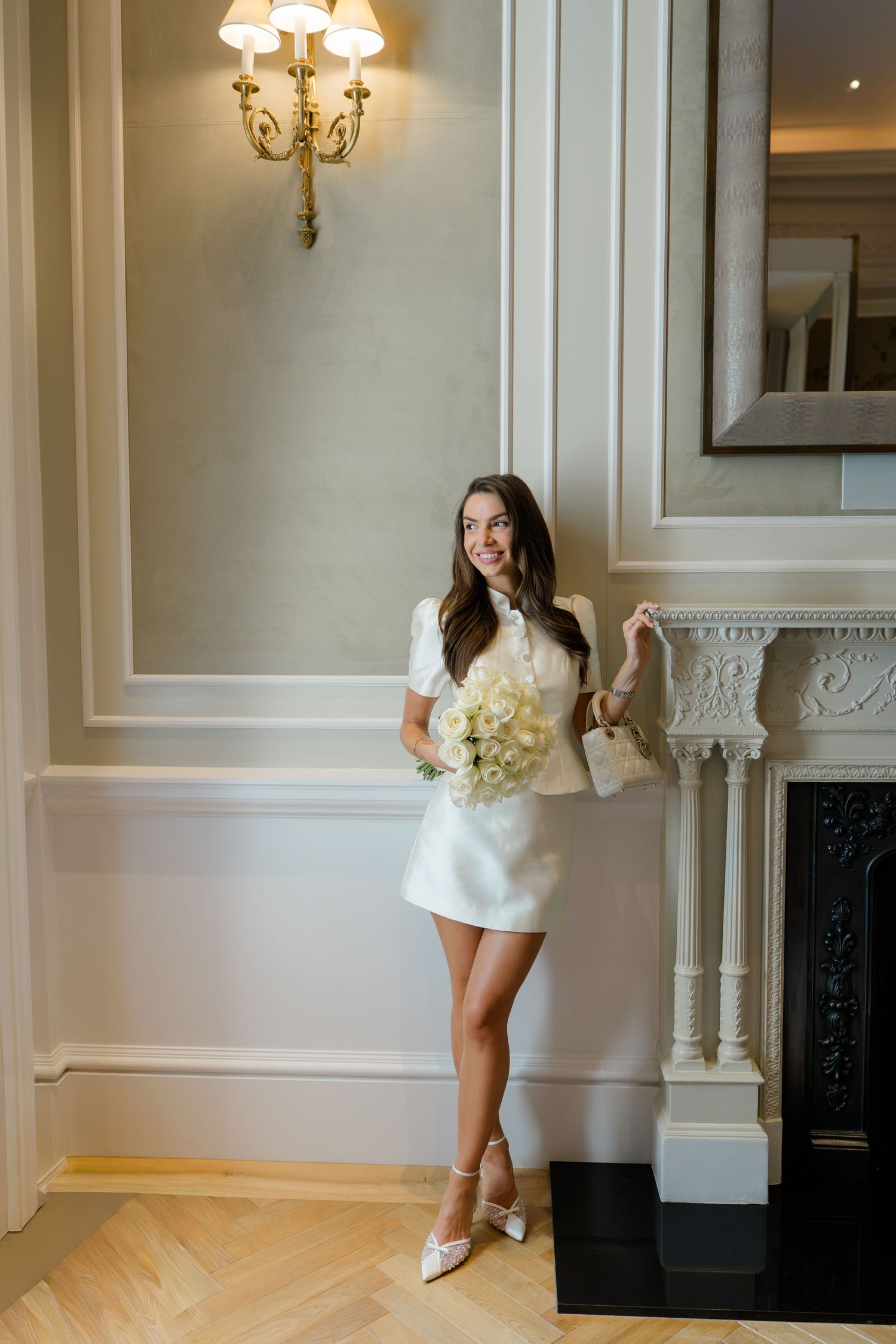 Smiling bride in short white dress holds bouquet beside elegant fireplace in classic interior.