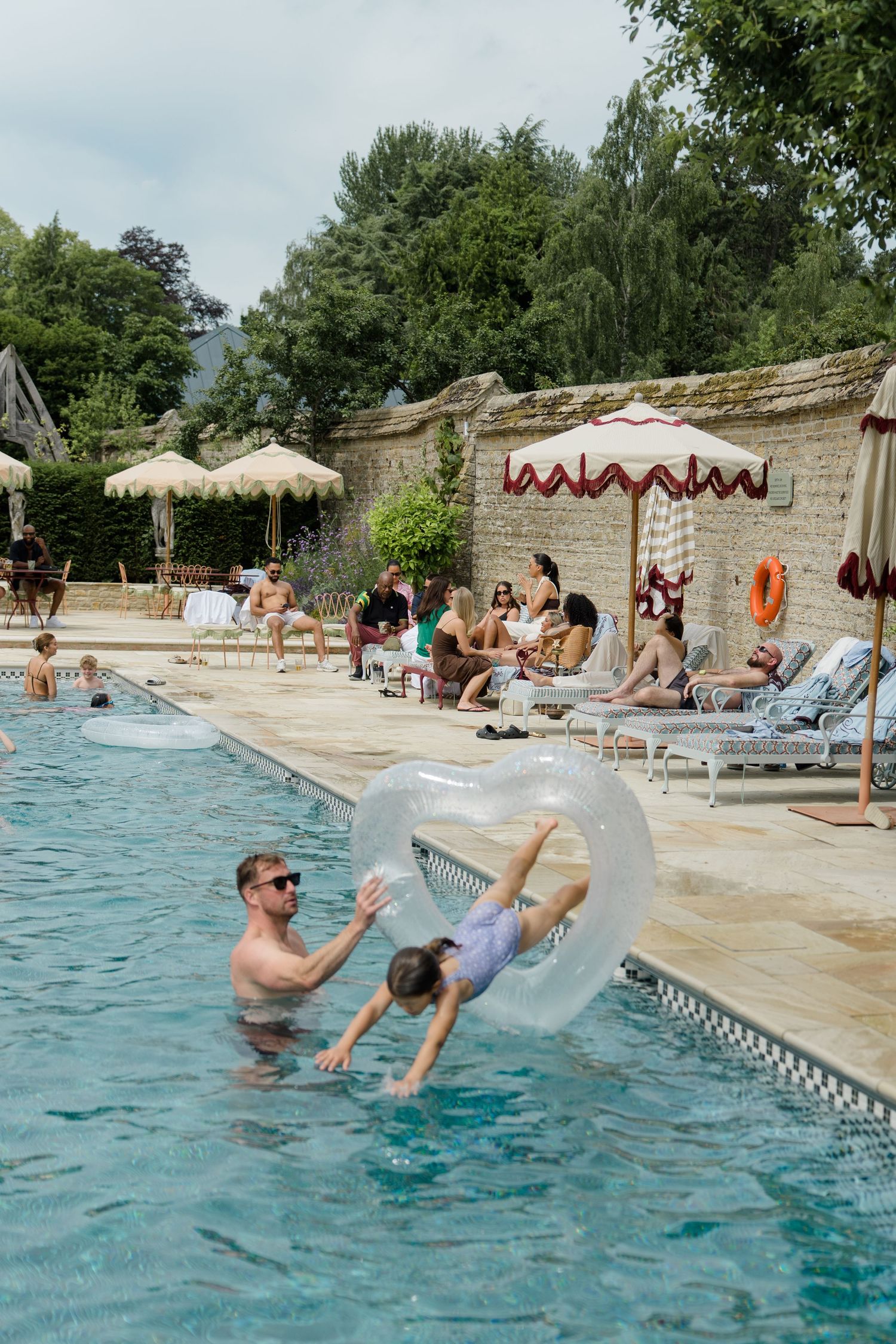 Wedding guests enjoy poolside celebration as man flips child through inflatable ring in sunny outdoor pool.