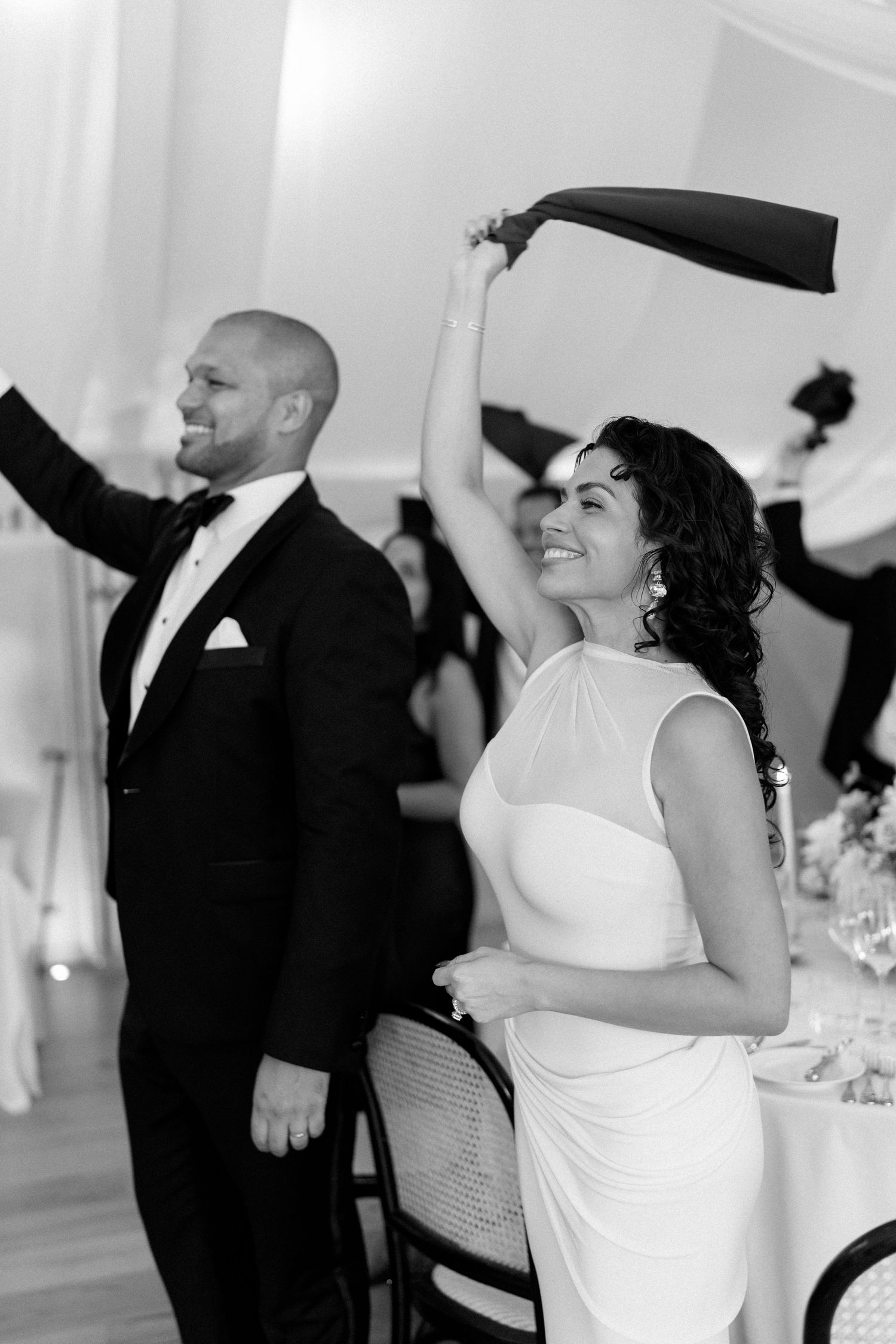 Black and white photo of joyful bride and groom waving napkins during lively wedding reception celebration.