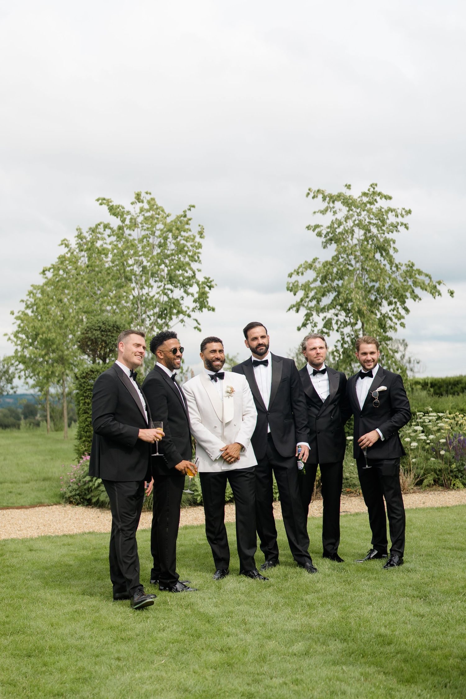 Groom in white tuxedo jacket laughing with groomsmen in black suits on manicured lawn.