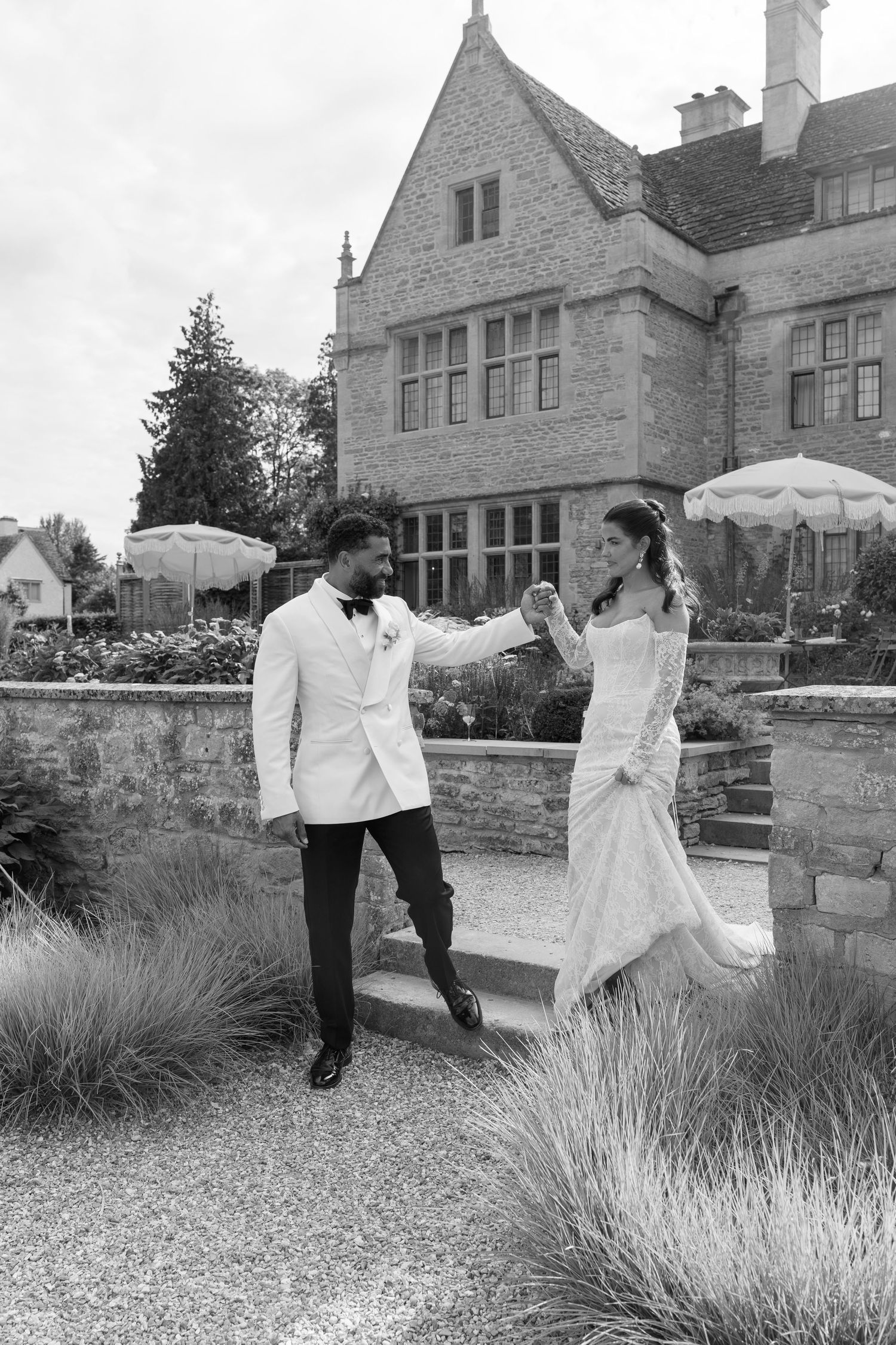 Bride and groom dance holding hands on garden steps of historic stone manor in black and white photo.