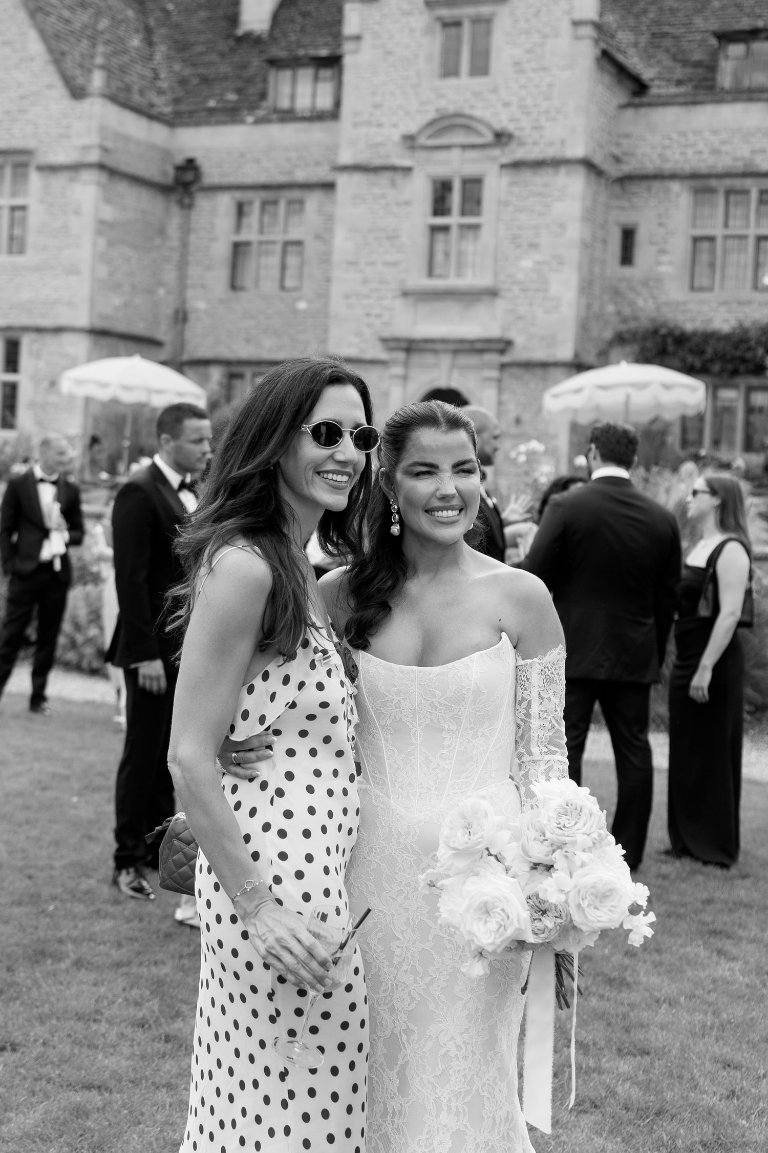 Bride in lace gown holds bouquet posing with guest in polka dot dress at elegant manor wedding reception.